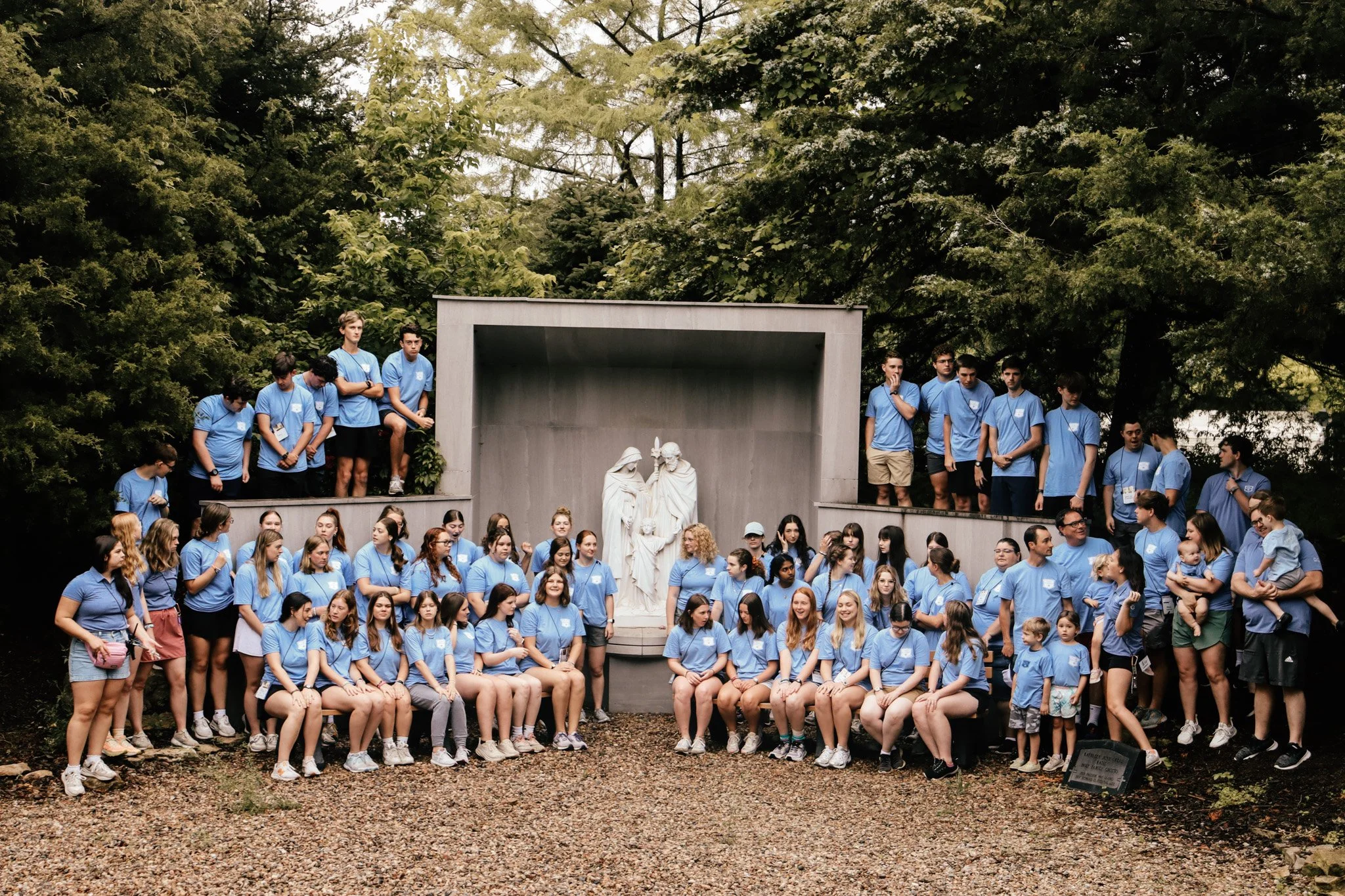 Large group of people, mostly young women, wearing matching blue t-shirts, gathered outdoors on a stone-paved area in front of a monument situated in a park with tall trees in the background.