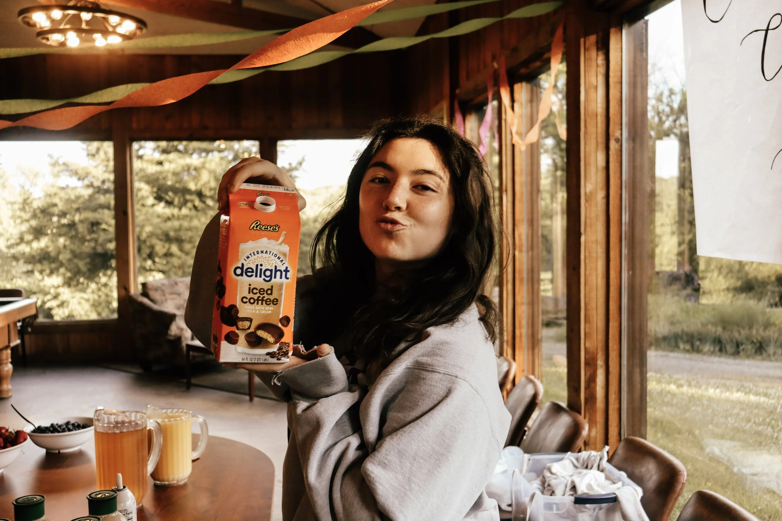 A woman with long dark hair holding a box of Reese's International Delight iced coffee, sitting at a table with glasses of orange-colored drinks and bowls of berries, inside a wooden room with large windows and trees outside.