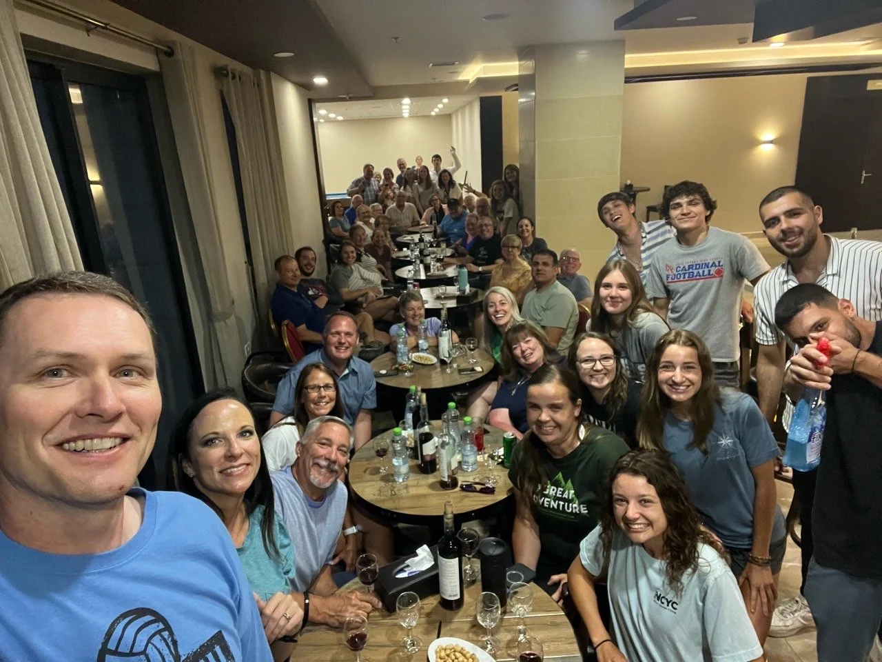 Group of people at a social gathering in a restaurant or event space, smiling and posing for a group selfie at a table with drinks and snacks.