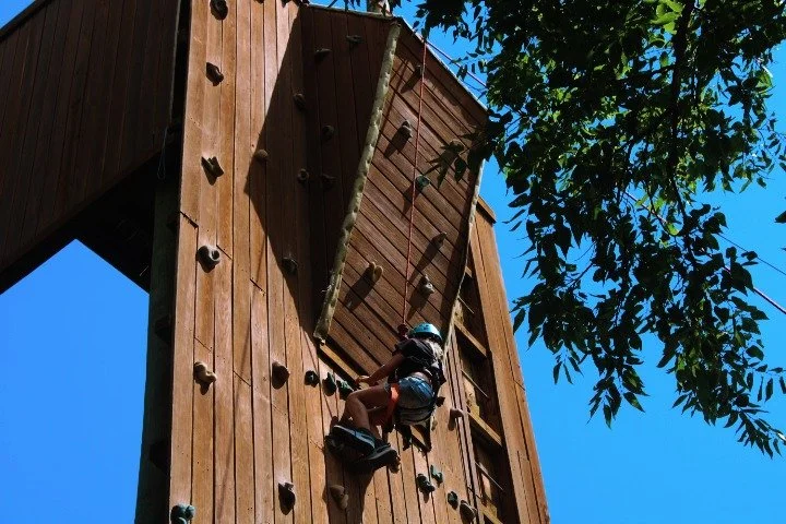 Child climbing a wooden outdoor rock climbing wall with harness and helmet on a sunny day, with trees and a blue sky in the background.