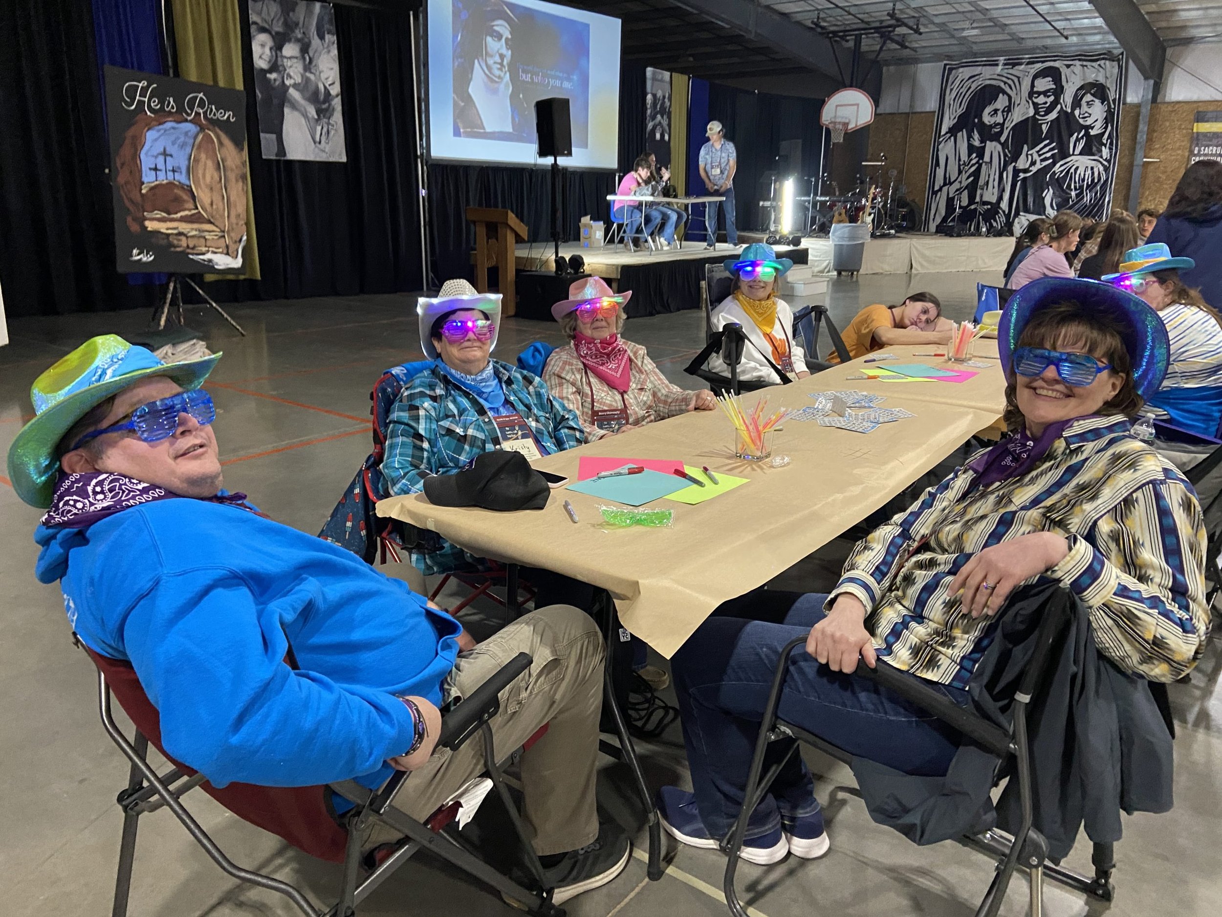 A group of seniors wearing colorful cowboy hats and sunglasses are sitting at a table in a gymnasium, attending an event with a stage and audience. The stage has a large screen displaying a woman speaking, and there are musical instruments and artwor
