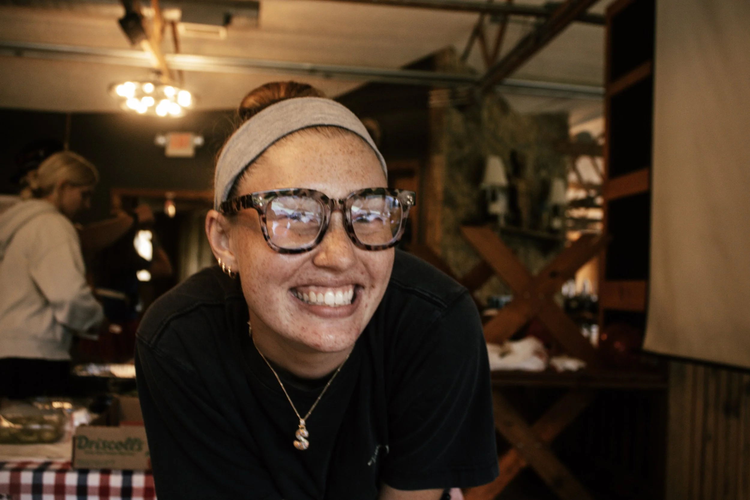 A smiling woman with freckles, wearing glasses, a gray headband, and a black shirt, sitting indoors at a social gathering or restaurant. In the background, there is another woman with blonde hair, glasses, and a beige jacket, near a wooden staircase 