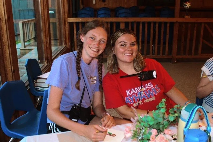 Two smiling young women sitting at a table indoors, with one wearing a Kansas Jayhawks t-shirt and the other in a light purple shirt. They are in a room with wooden walls, and there are blue chairs and some plants on the table.