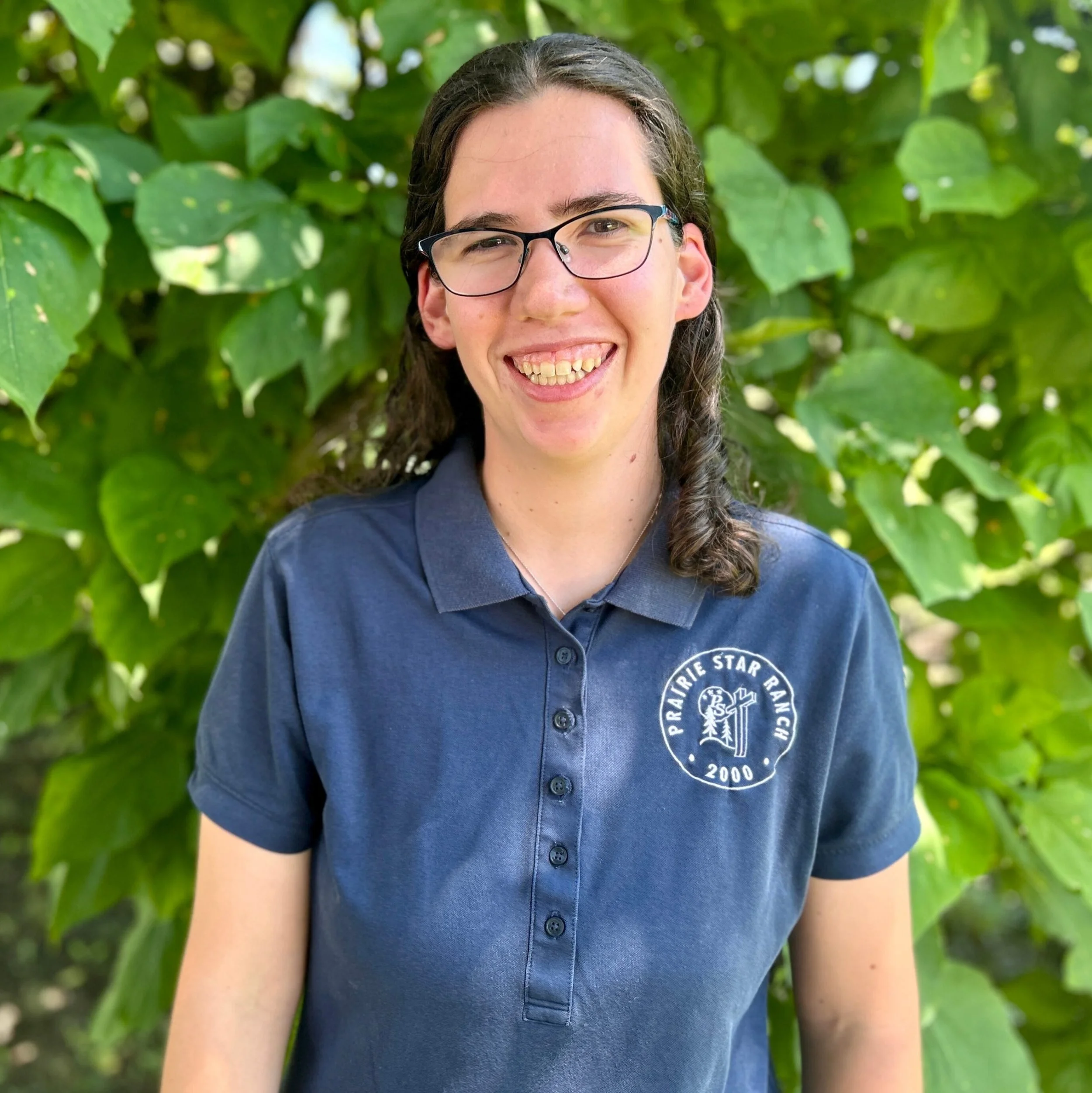 Close-up of a person with long dark hair, wearing glasses and a navy blue polo shirt, smiling outdoors in front of green leafy background.