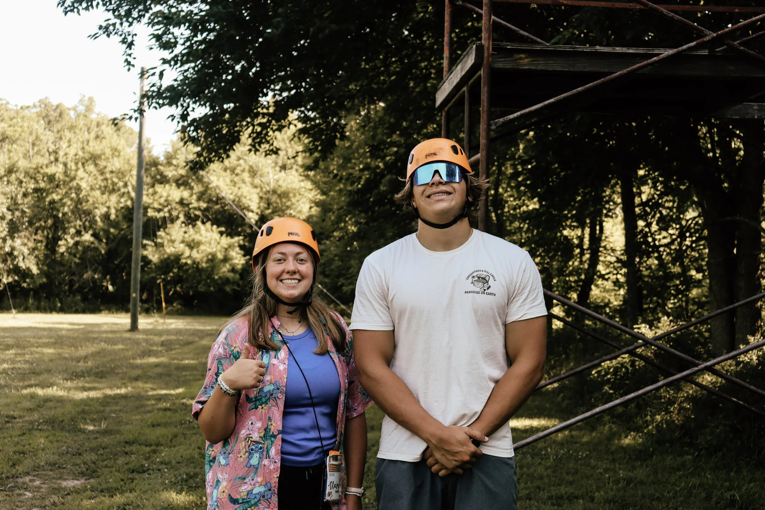 Two young people wearing orange helmets and safety gear, smiling outdoors in a park with trees and a metal structure in the background.