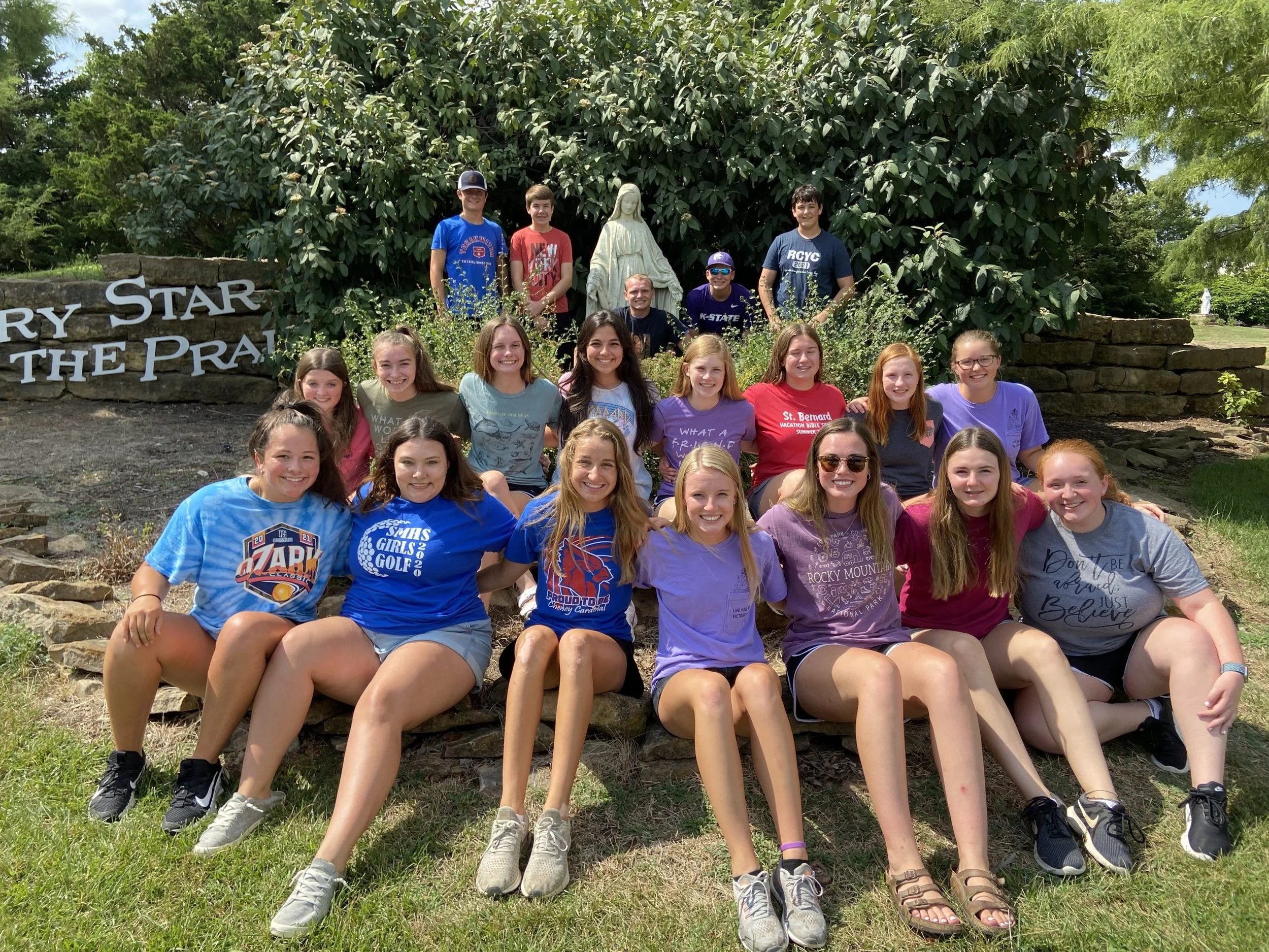 Group of smiling teenagers sitting on stone stairs in front of a large green bush and a statue, with a sign that reads 'St. Bernad'.
