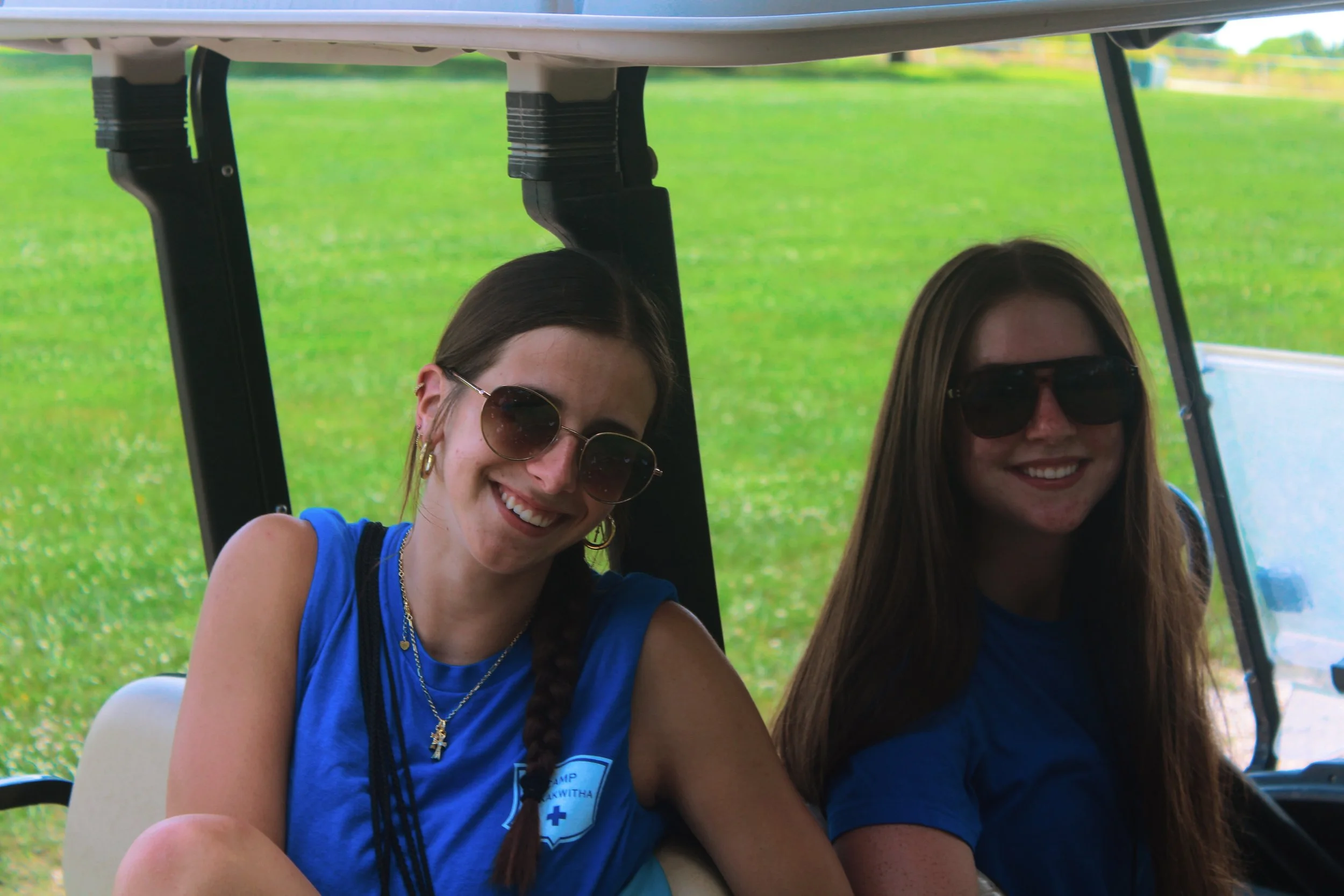 Two women with long hair and sunglasses smiling while sitting in a golf cart on a grassy field.