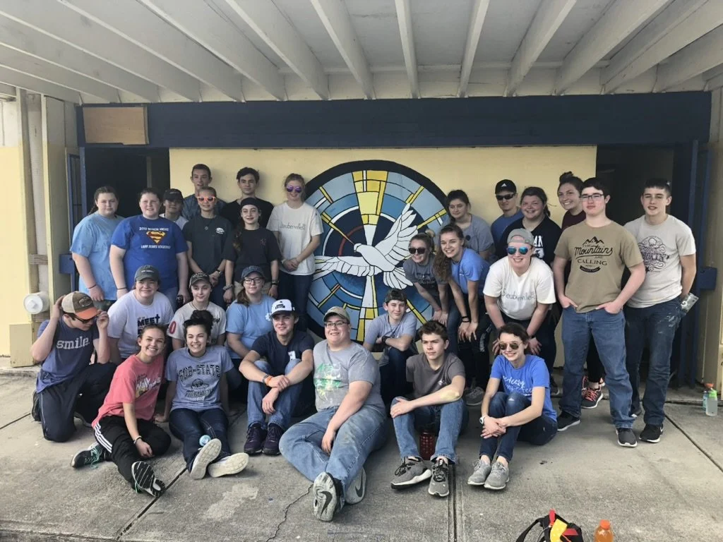 Group of teenagers posing in front of a mural with a dove and a clock on a yellow wall.