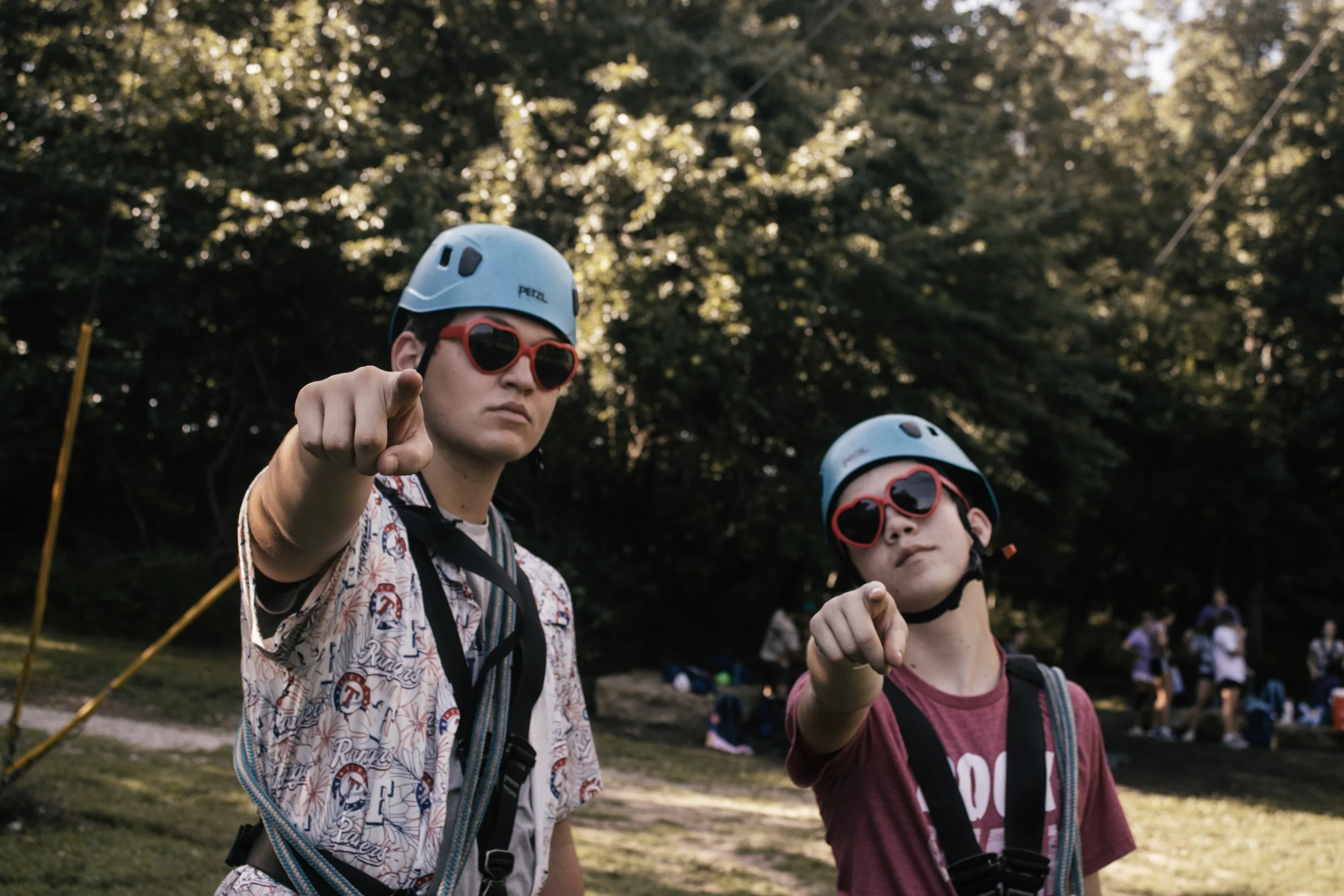 Two individuals wearing helmets and sunglasses pointing at the camera during an outdoor event in a park, with trees and other people in the background.