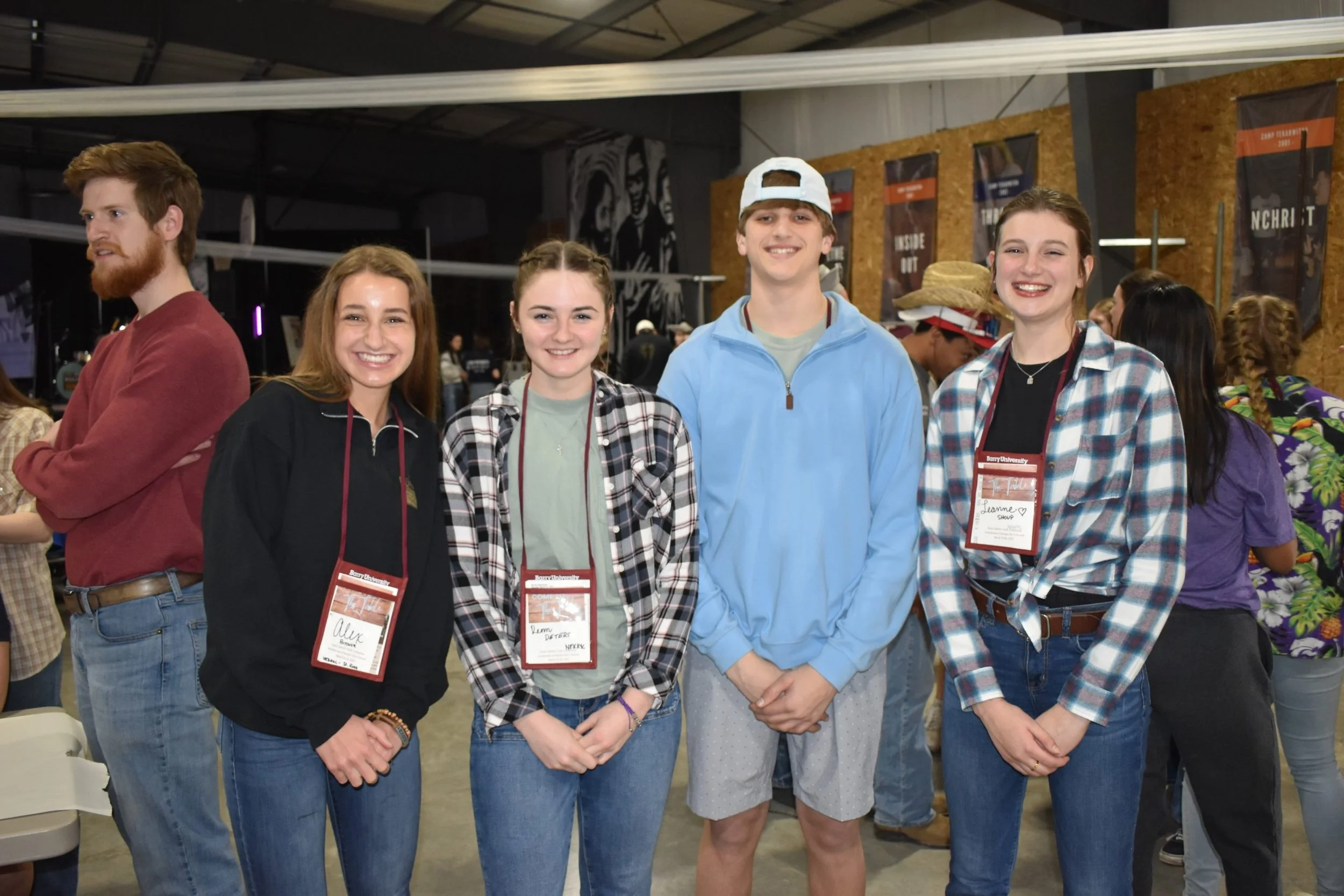 Group of five young adults standing together indoors, smiling, with some wearing name tags, at a social or university event.
