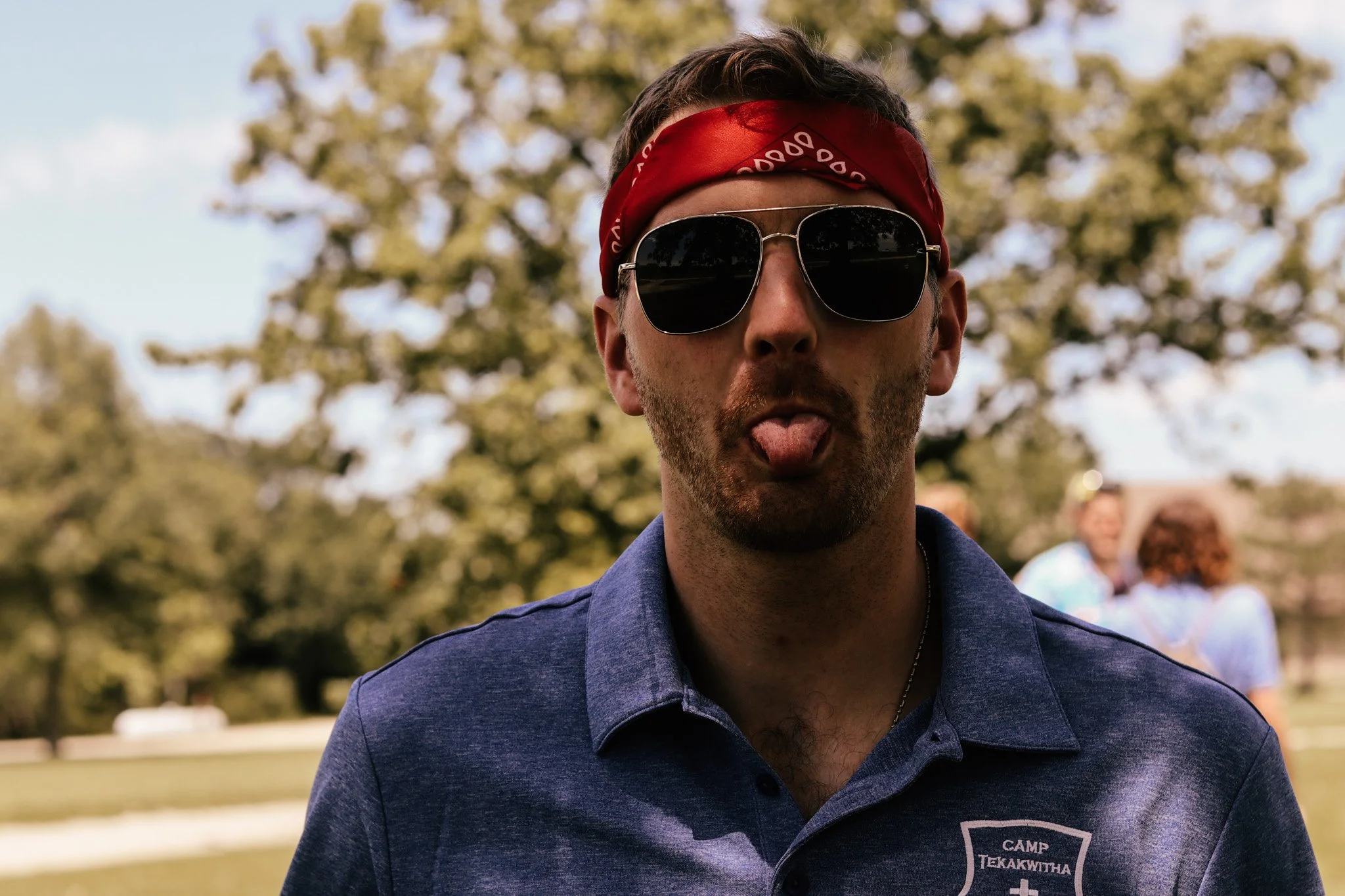 A young man wearing sunglasses and a red bandana sticking out his tongue outdoors with a blurred background of trees and people.