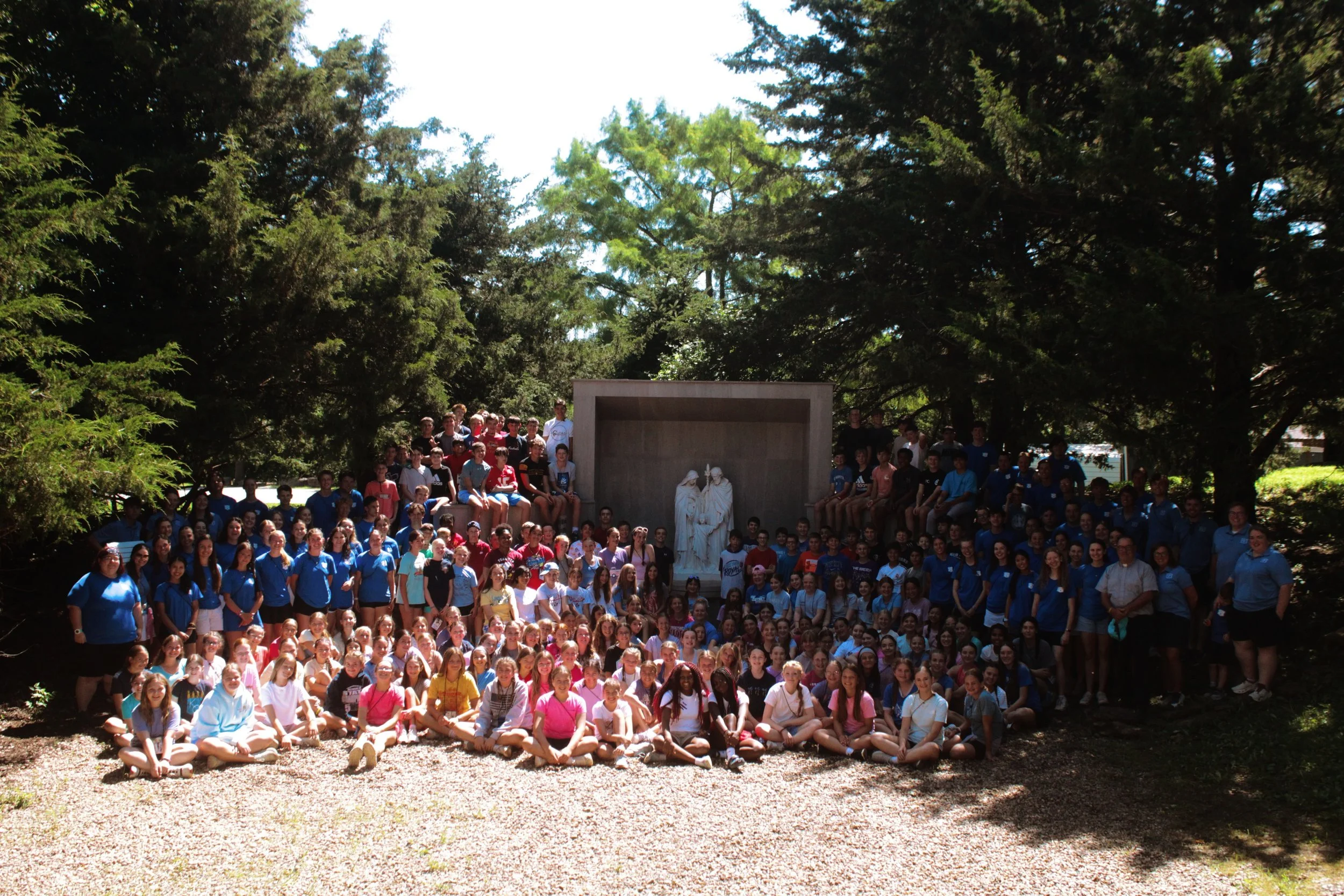 A large group of children and teenagers posing outdoors in front of a sculpture of Jesus and two other figures, surrounded by trees and greenery.