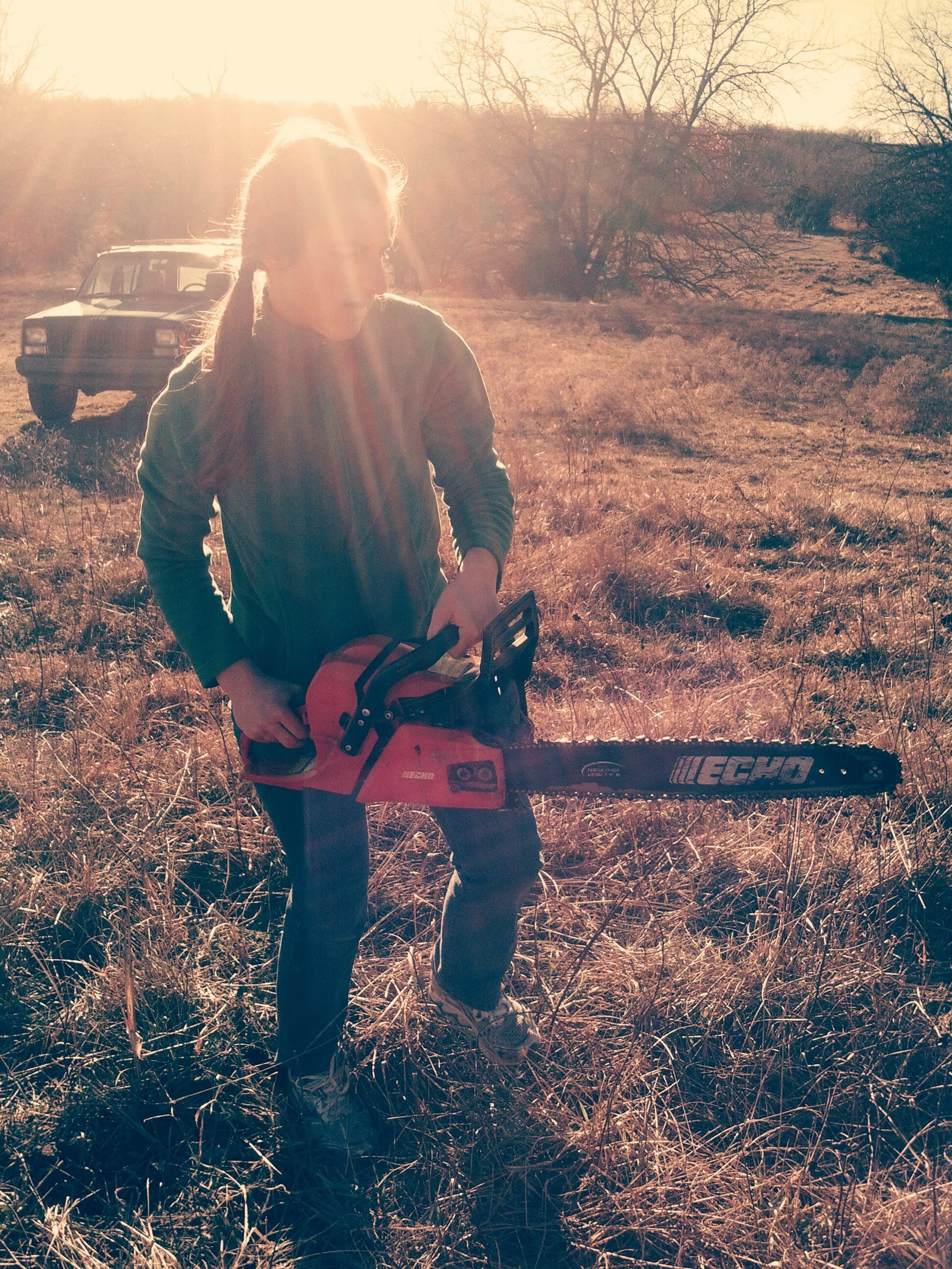 A woman carying a chainsaw in a meadow, with a bright sunset behind her.