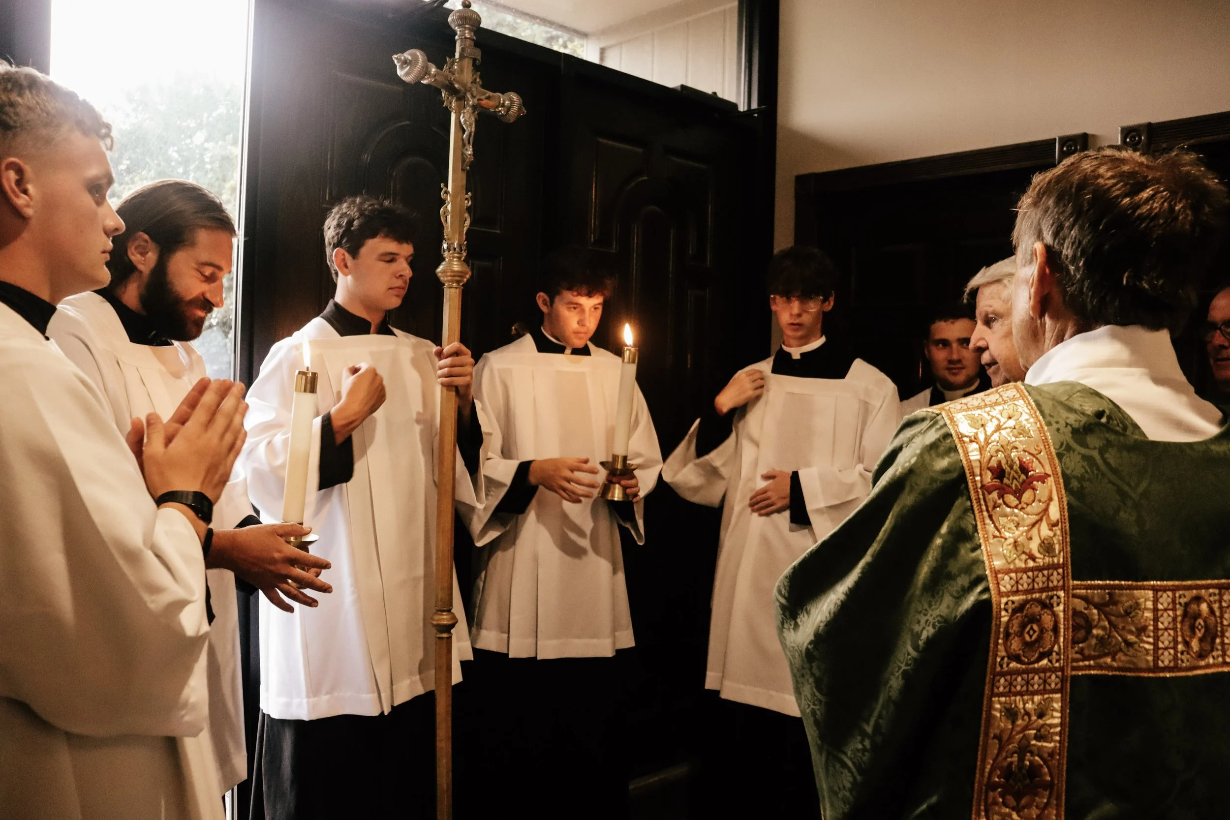 A religious ceremony taking place with an individual holding a crucifix and a lit candle, surrounded by people dressed in white robes, possibly priests or altar servers, in a church setting.