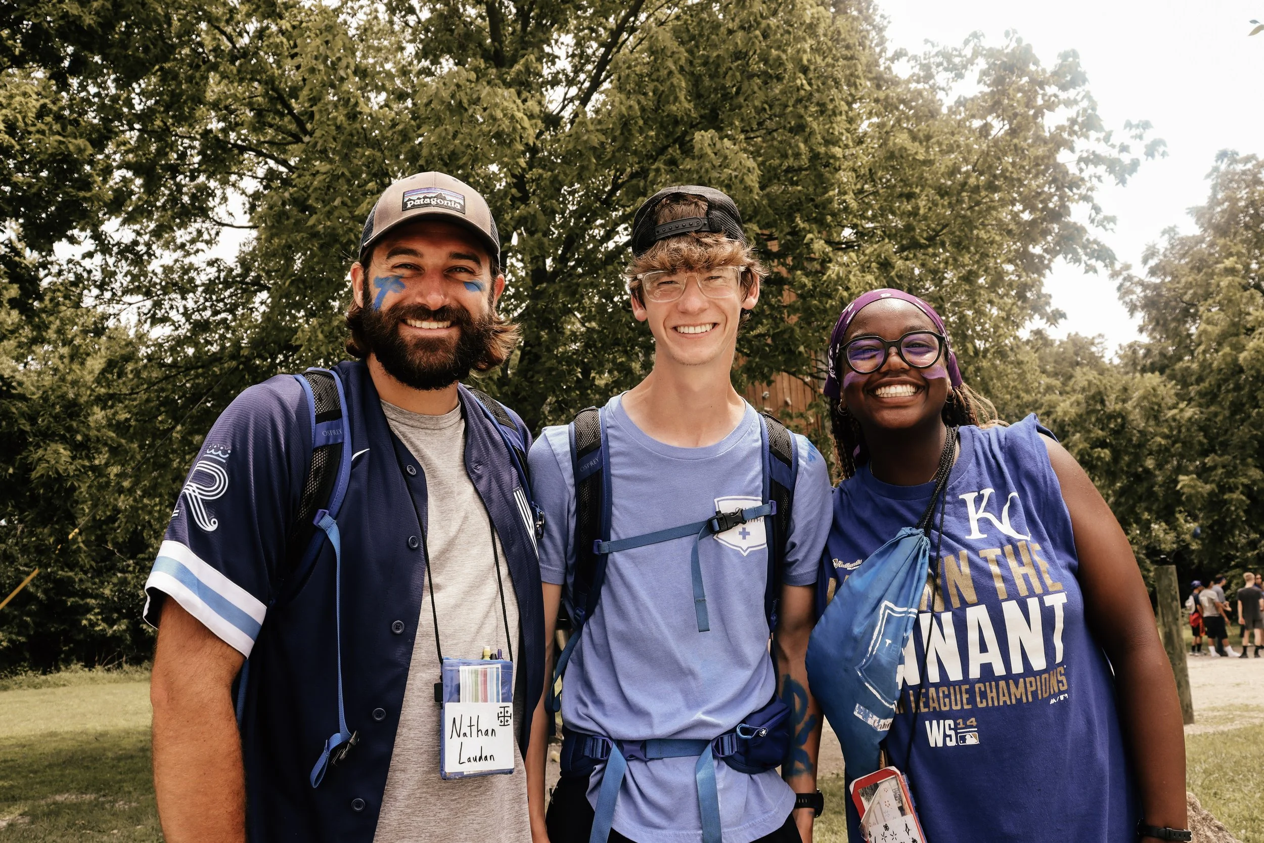 Three young adults standing outdoors in front of trees, smiling. The person on the left has a beard, is wearing a baseball cap, a blue sports jersey, and has face paint and a name tag that reads 'Nathan Laudan.' The person in the middle has glasses, 