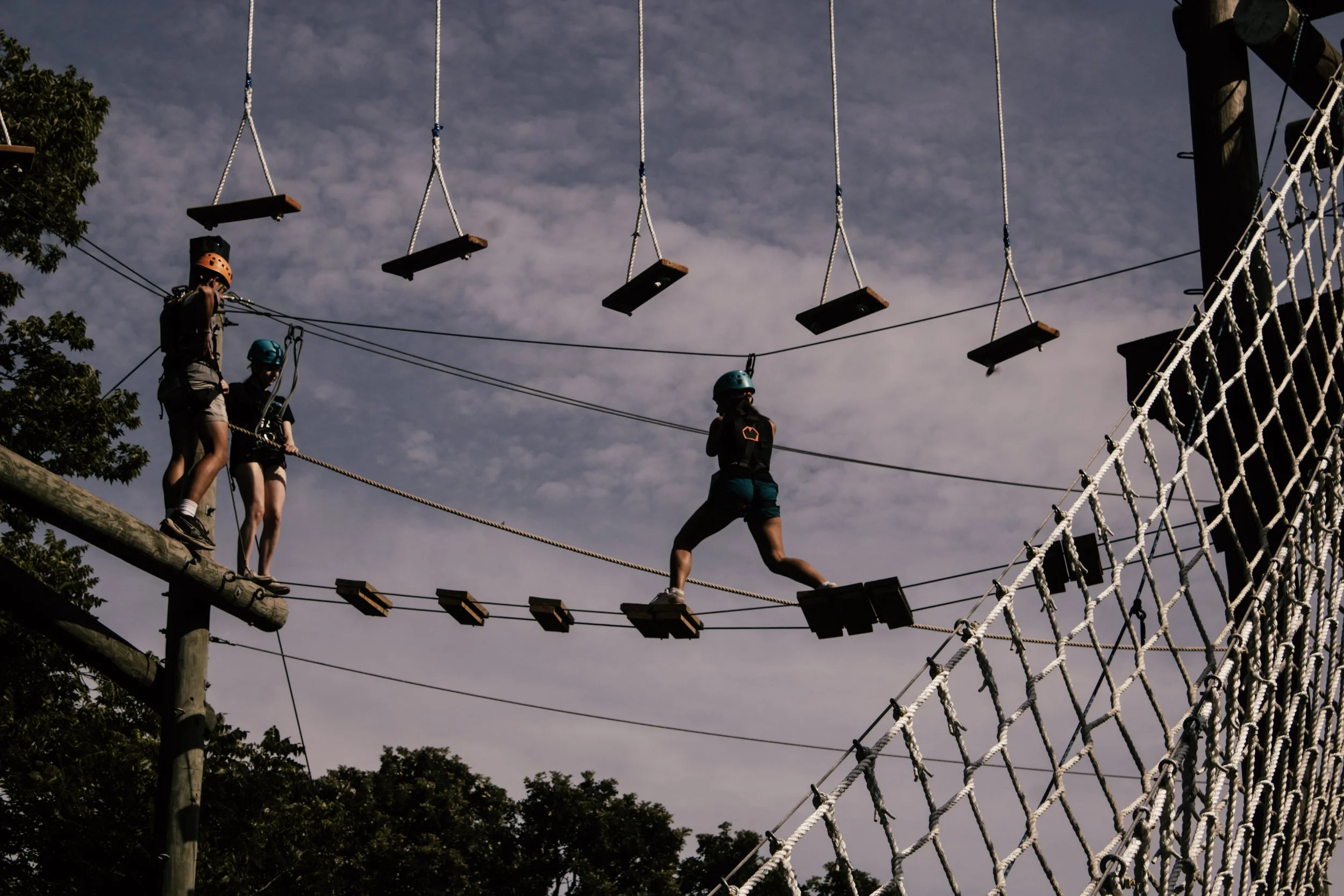 People wearing helmets and harnesses crossing a ropes course with swings and a netted obstacle, outdoors against a cloudy sky.