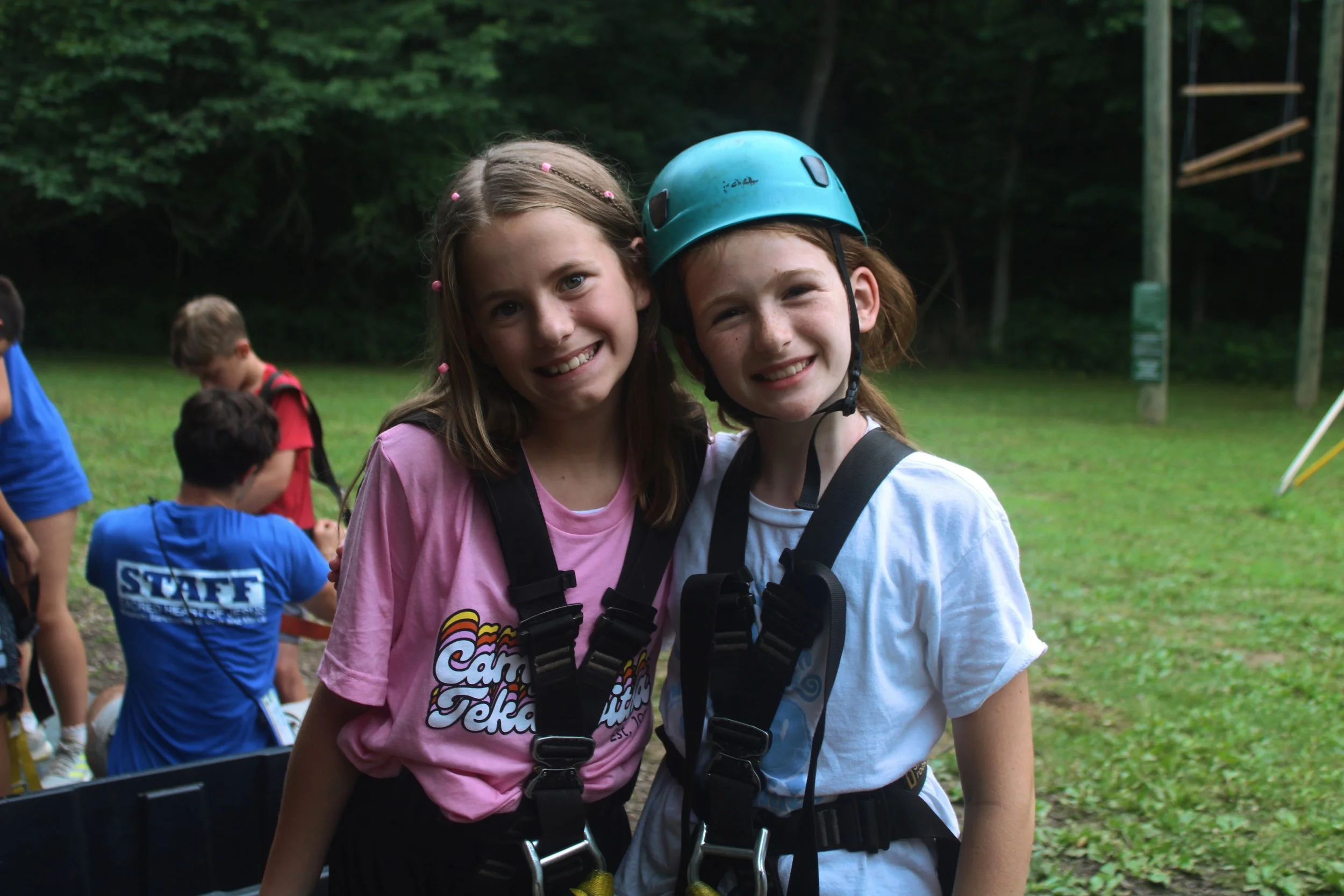 Two young girls smiling, standing outdoors, wearing helmets and harnesses, with a group of children in the background at a park or playground.