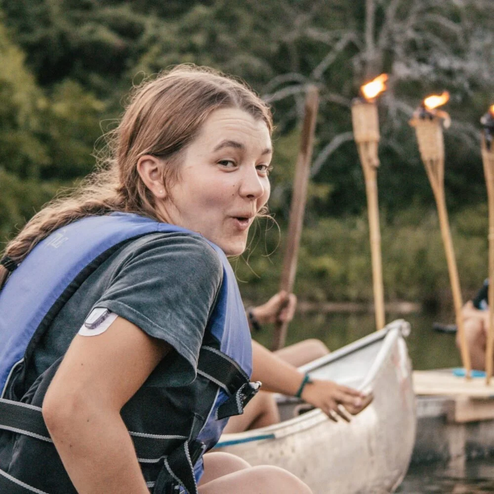 A young woman with long braided hair wearing a life jacket and gray t-shirt, sitting near a canoe on a river, surrounded by trees, with tiki torches in the background.