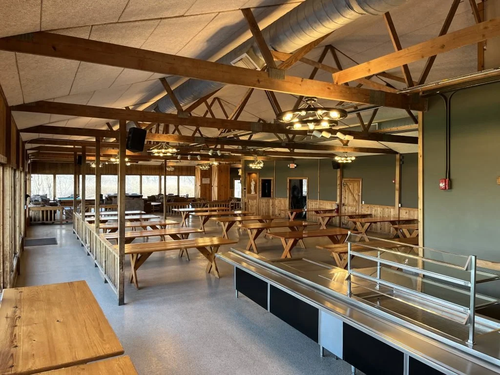 Empty rustic dining hall with wooden picnic tables, green walls, and large windows, featuring a serving counter and ceiling fans.