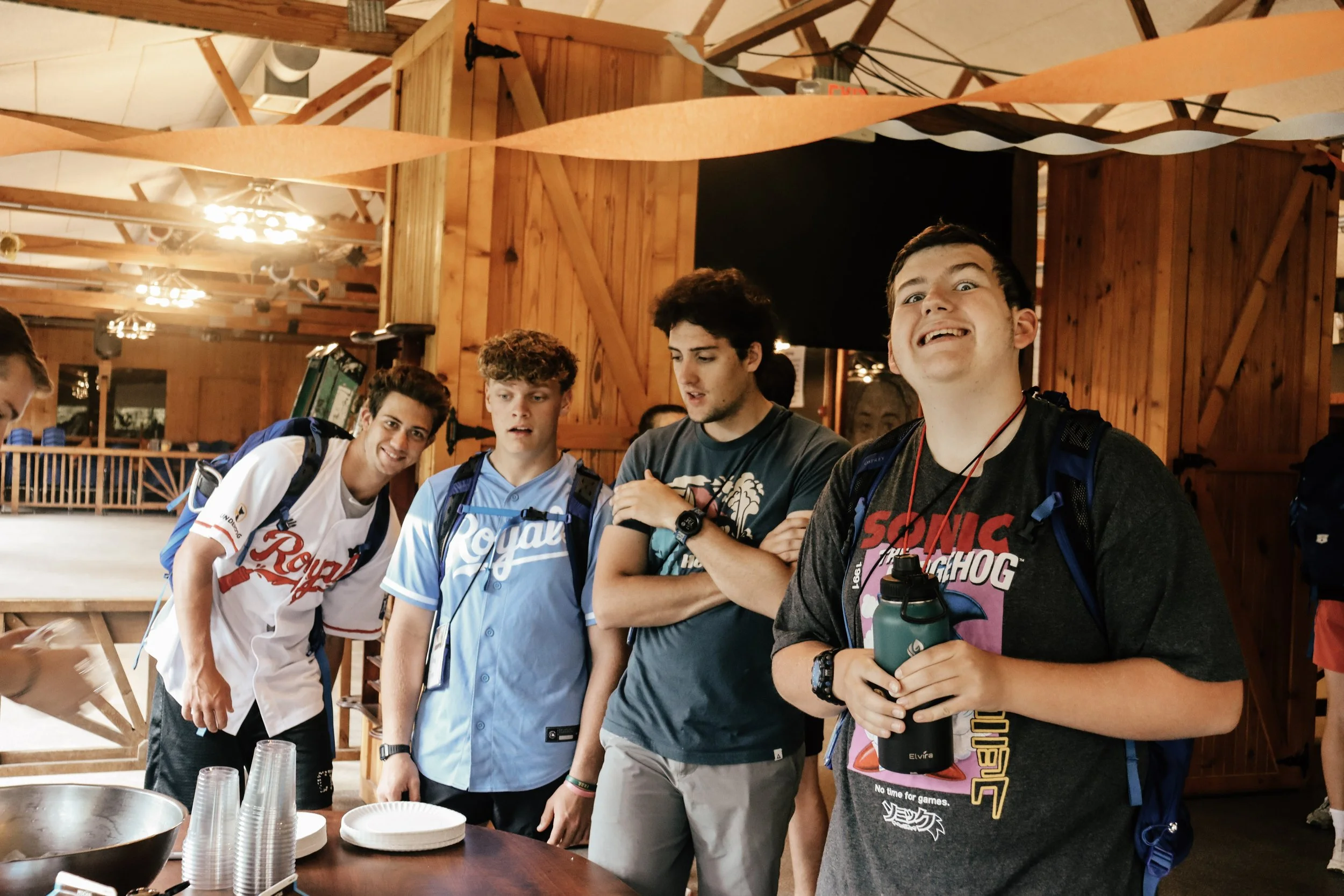 A group of young men standing together in a rustic wooden hall, engaged in conversation. They are dressed casually, some wearing backpacks and holding water bottles, and appear cheerful and relaxed.