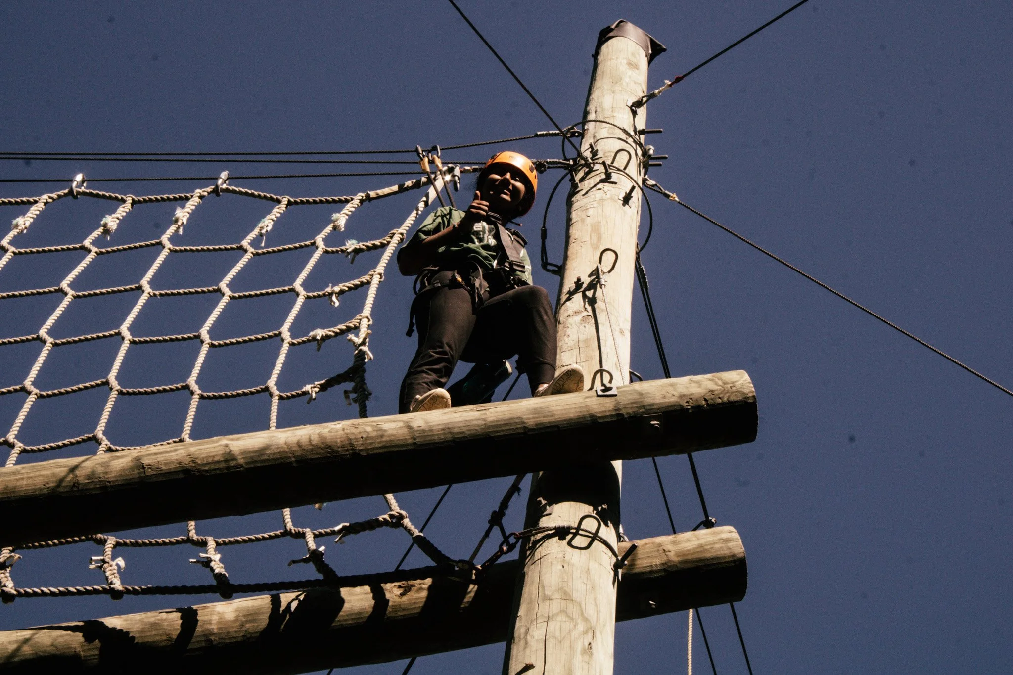 A young boy wearing a helmet and harness, climbing a wooden and rope obstacle on a high ropes course against a clear blue sky.