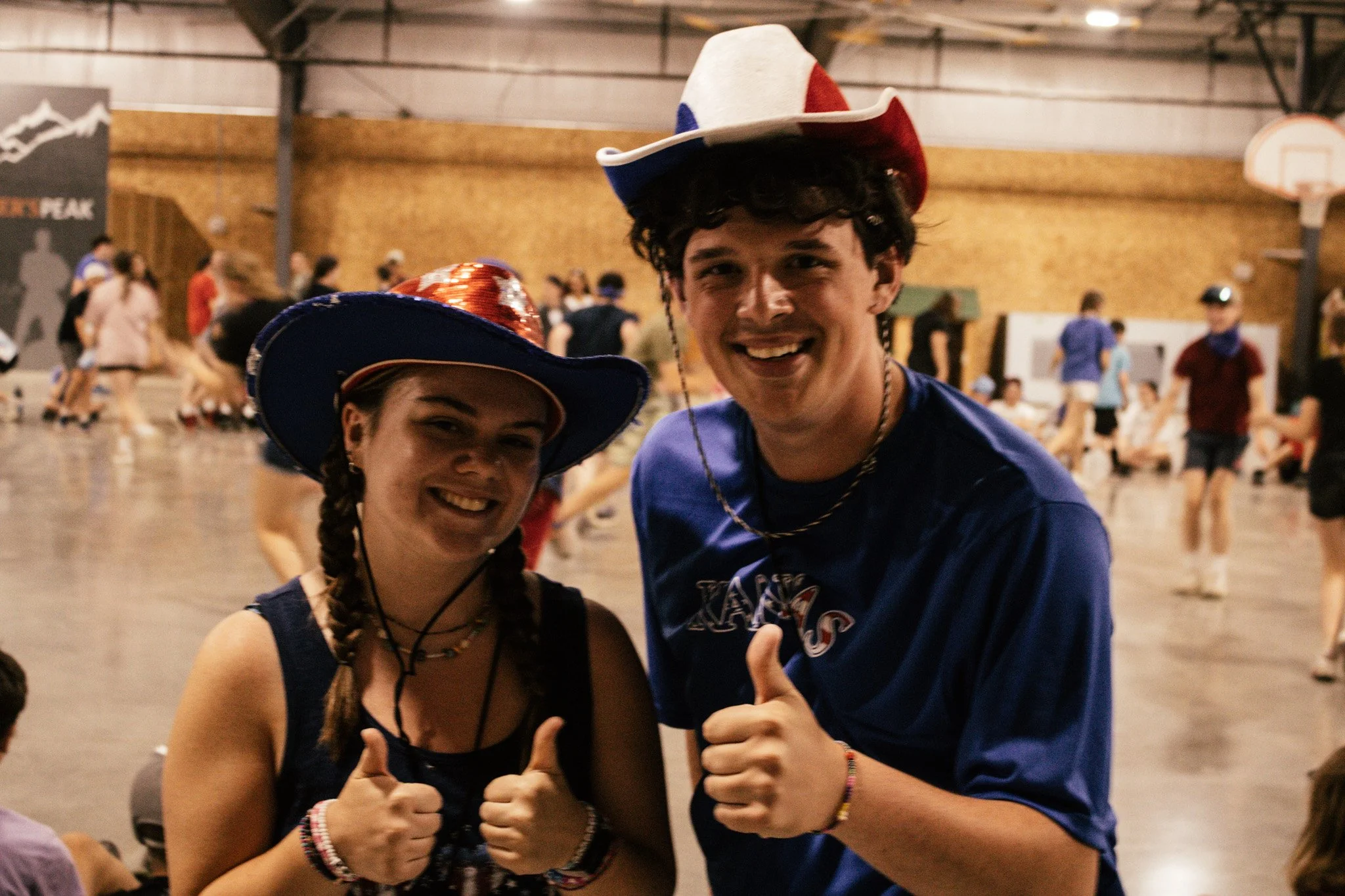 Two young people wearing patriotic hats smiling and giving thumbs up at an indoor roller skating rink filled with other skaters in the background.