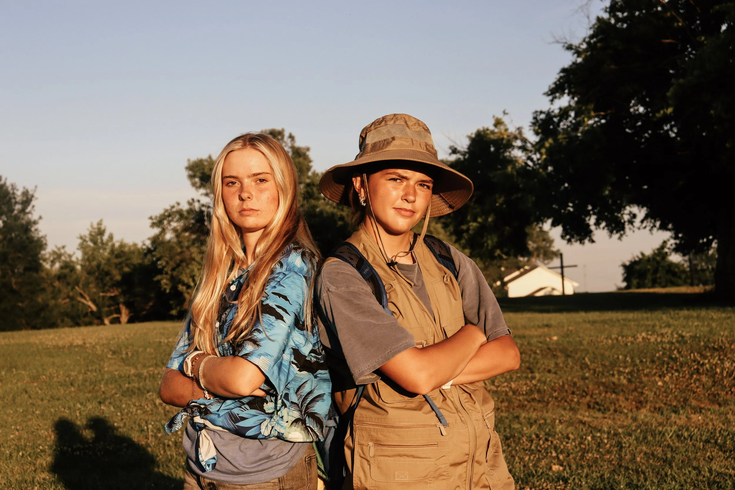 Two young women stand outdoors with arms crossed. One has long blonde hair and wears a blue tropical shirt; the other has short hair, a tan sun hat, and a tan vest. The background includes trees, a house, and a clear sky.