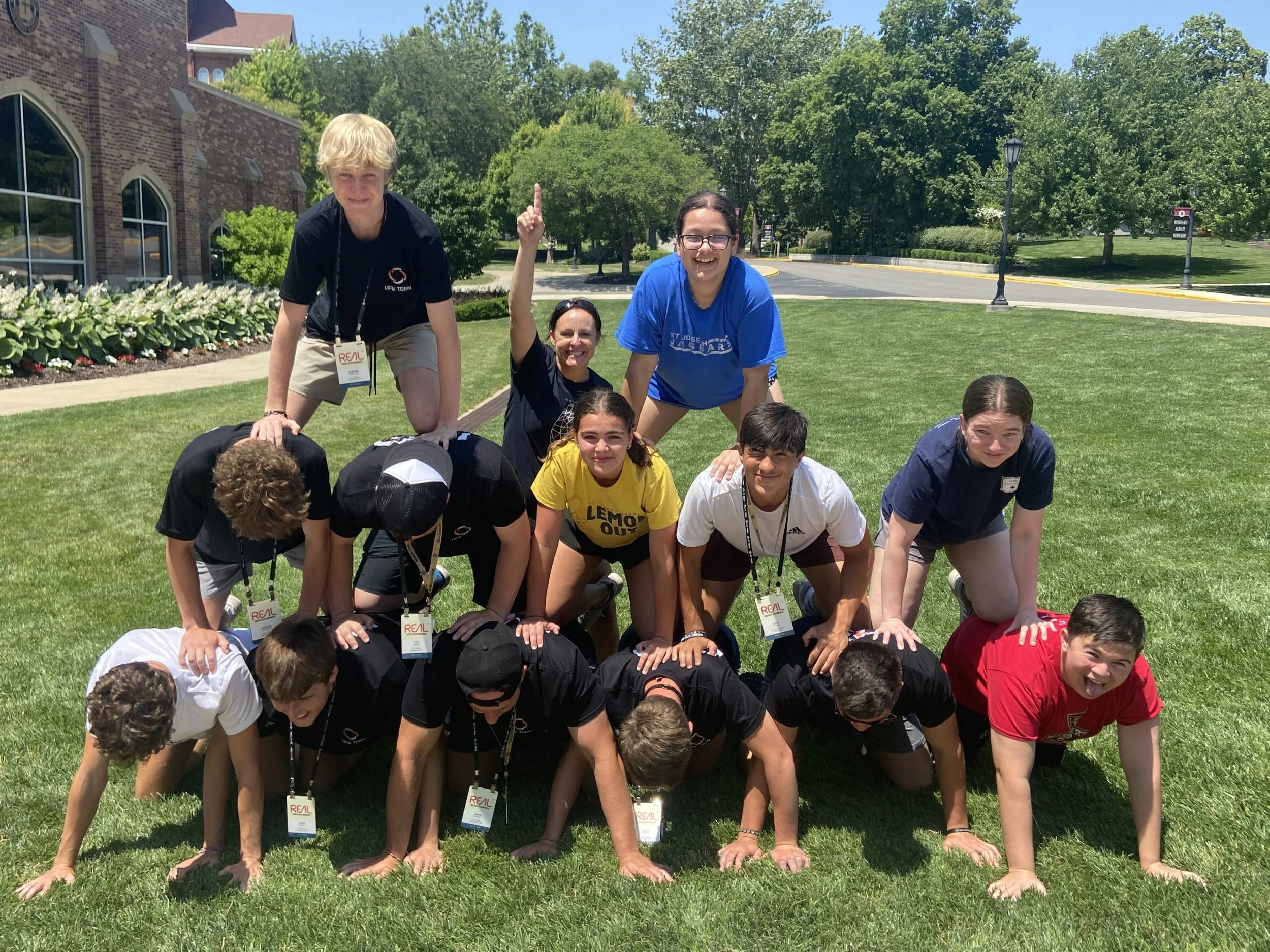 A group of children and a woman forming a human pyramid outdoors on a grassy area with trees and a brick building in the background on a sunny day.