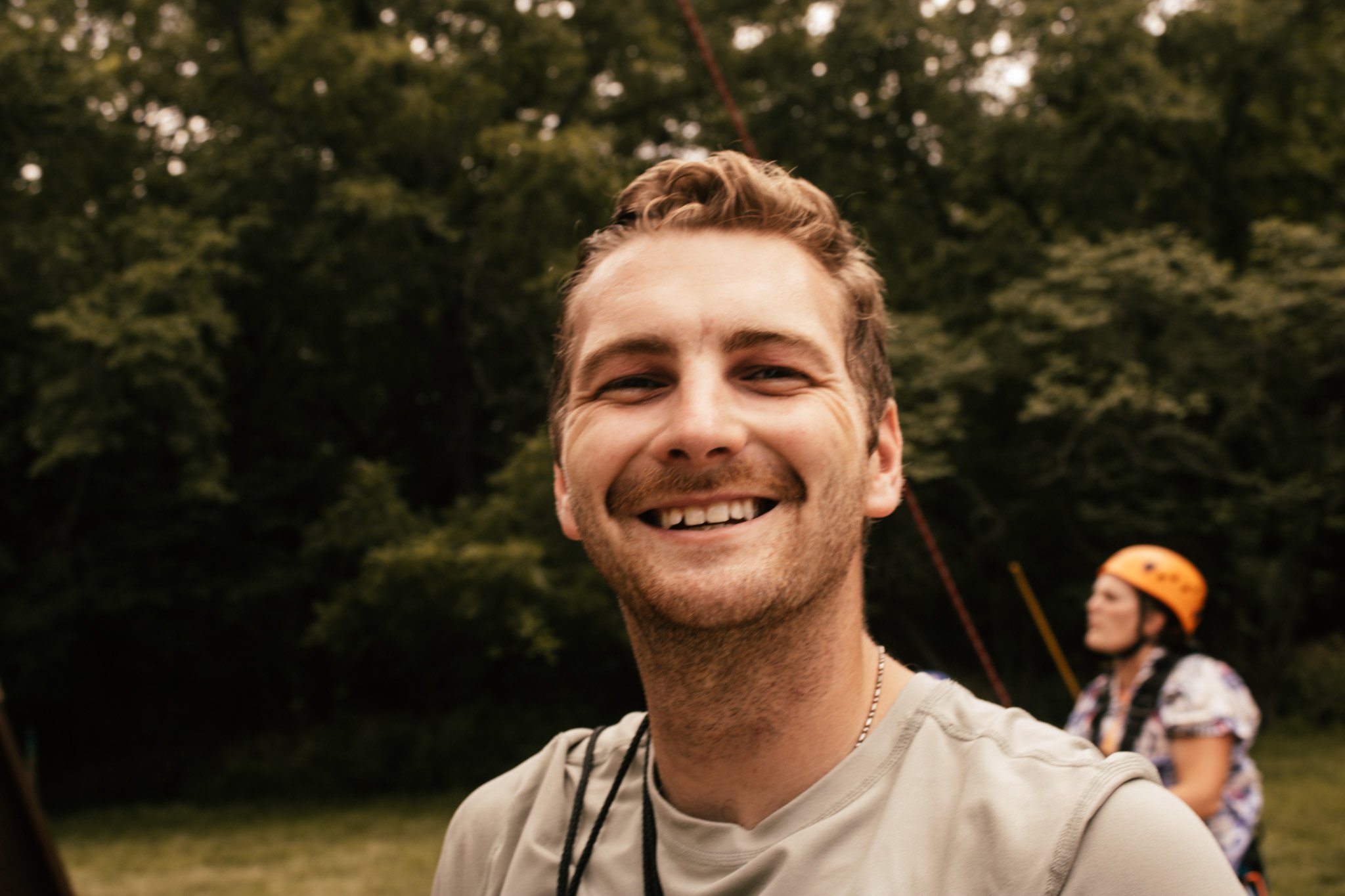 Close-up of a smiling young man with short light brown hair, wearing a light-colored shirt outdoors with trees in the background, during daytime.
