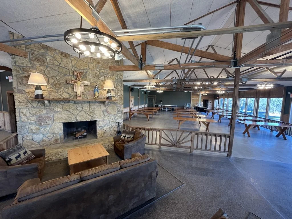 Lodge-style event hall with stone fireplace, wooden beams, and multiple picnic tables. Cozy seating area with couches and armchairs near the fireplace, and large windows along one side of the room.