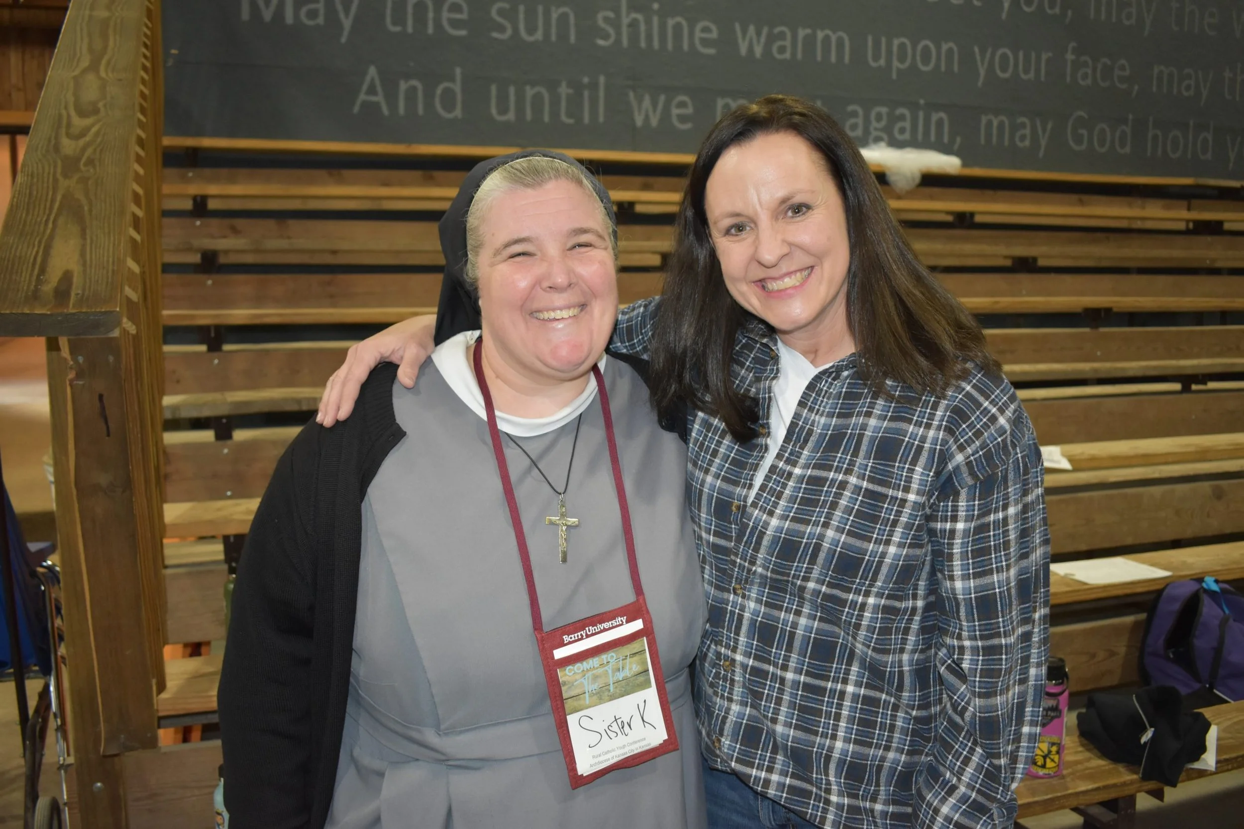 Two women smiling and standing close together at an indoor event, with wooden bleachers and a large sign with text in the background. One woman is wearing a black head covering and crosses necklace, while the other has long brown hair and is wearing 