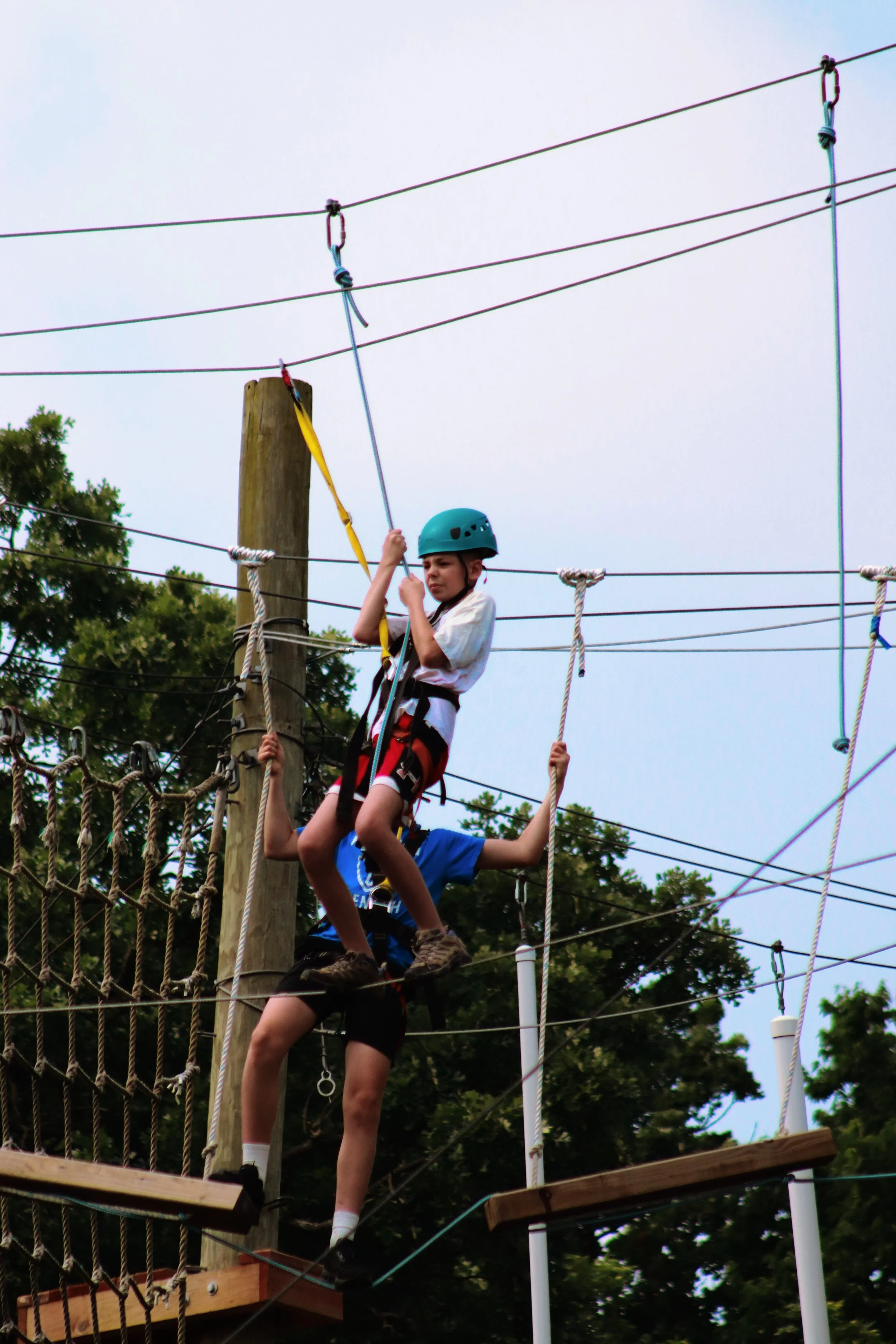 Two children, one in a blue helmet and another in a white shirt, are climbing a ropes course. The child in the blue helmet is holding a yellow safety line, and they are both on a wooden platform, surrounded by ropes and safety harnesses, with trees i