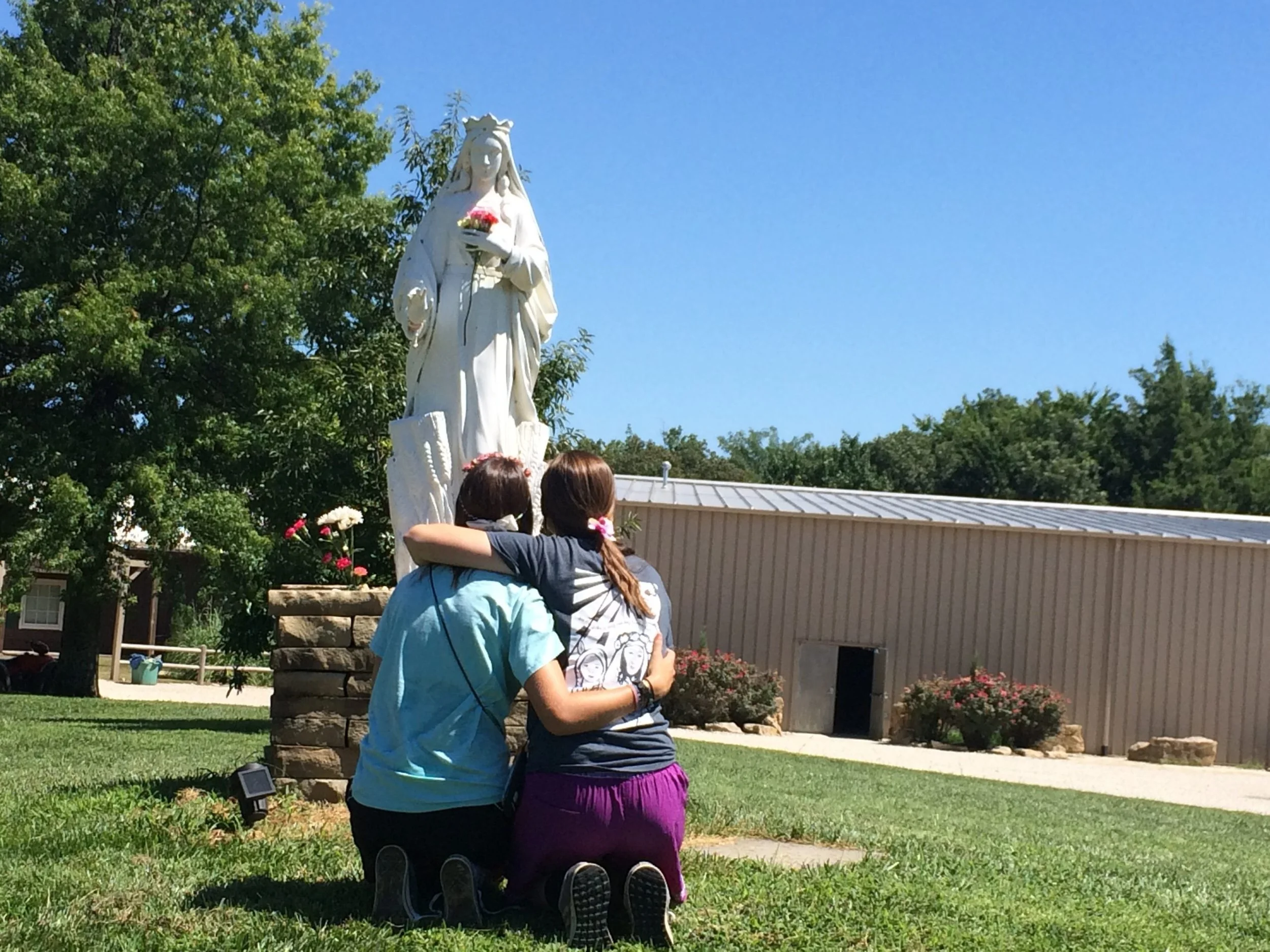 A young boy and girl kneel on the grass, hugging each other while facing a statue of the Virgin Mary holding a bouquet of flowers. The scene takes place outdoors on a sunny day with green trees and a building in the background.