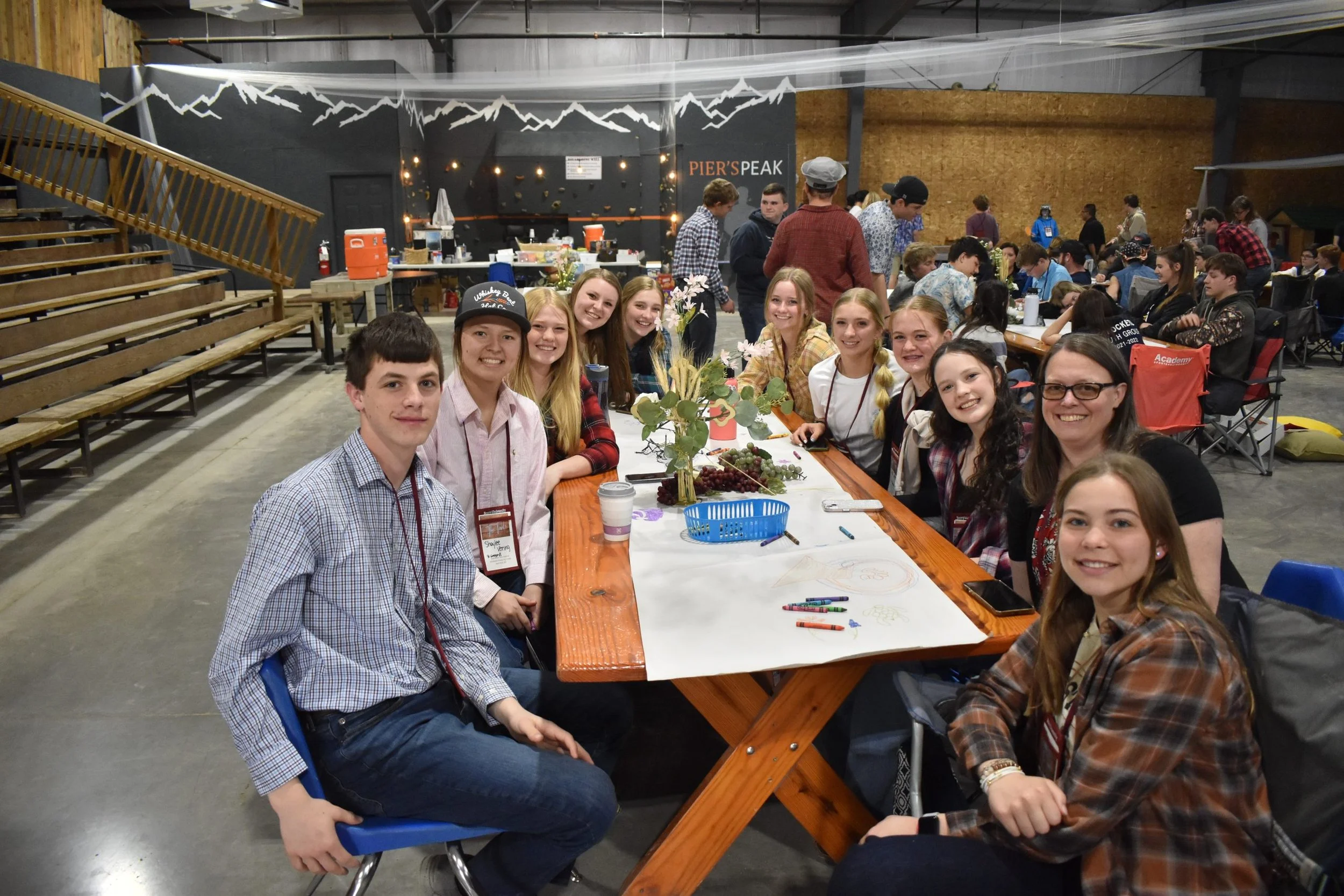 A group of young people sitting at a long table decorated with a small floral centerpiece, in a large indoor space with a mountain mural on the wall behind them. Many other people are standing or sitting in the background, and there is a table with f