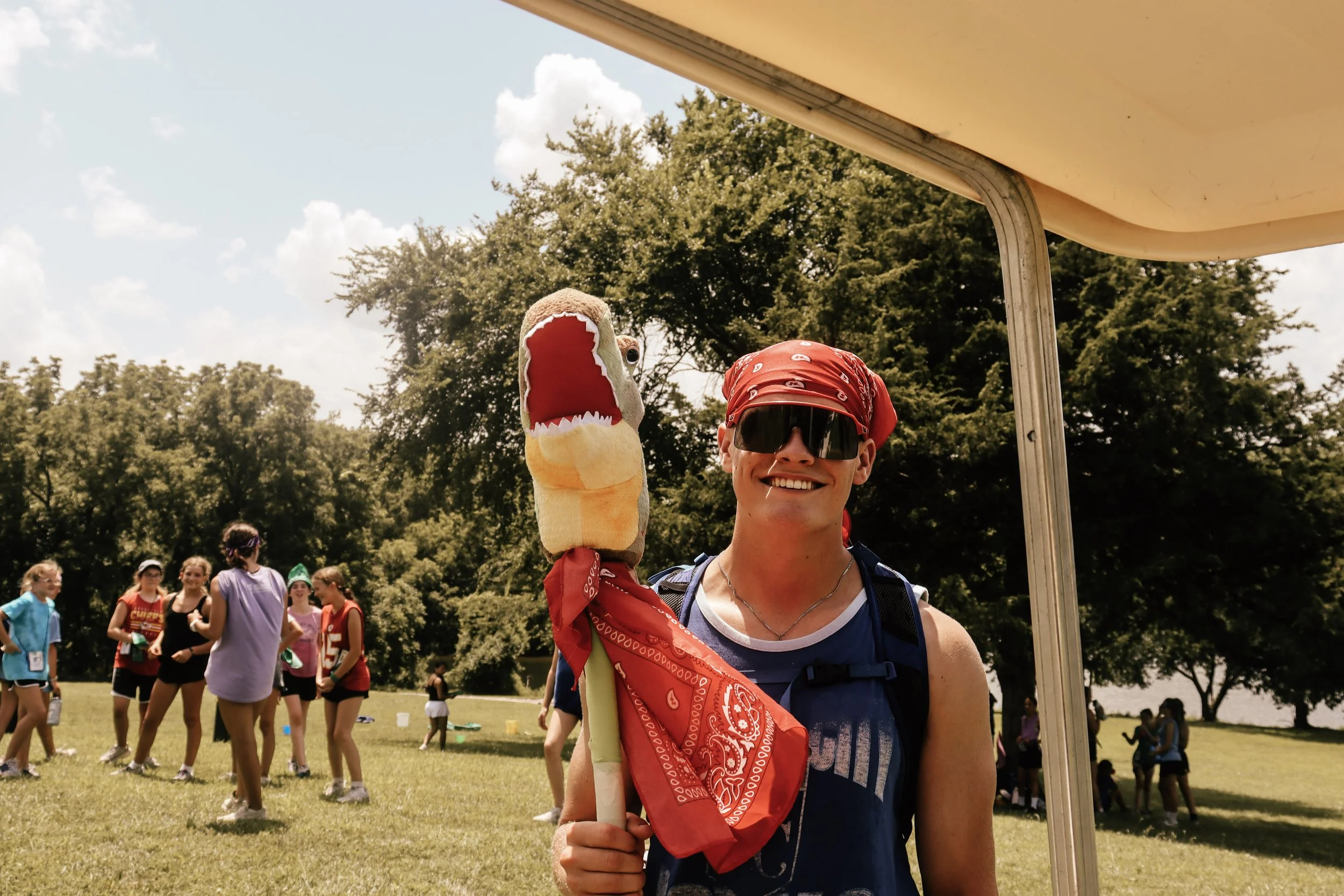 A young man smiling and wearing sunglasses, a red bandana, and a blue shirt, holding a plush dinosaur toy. In the background, a group of people is gathered in a park under a partly cloudy sky.