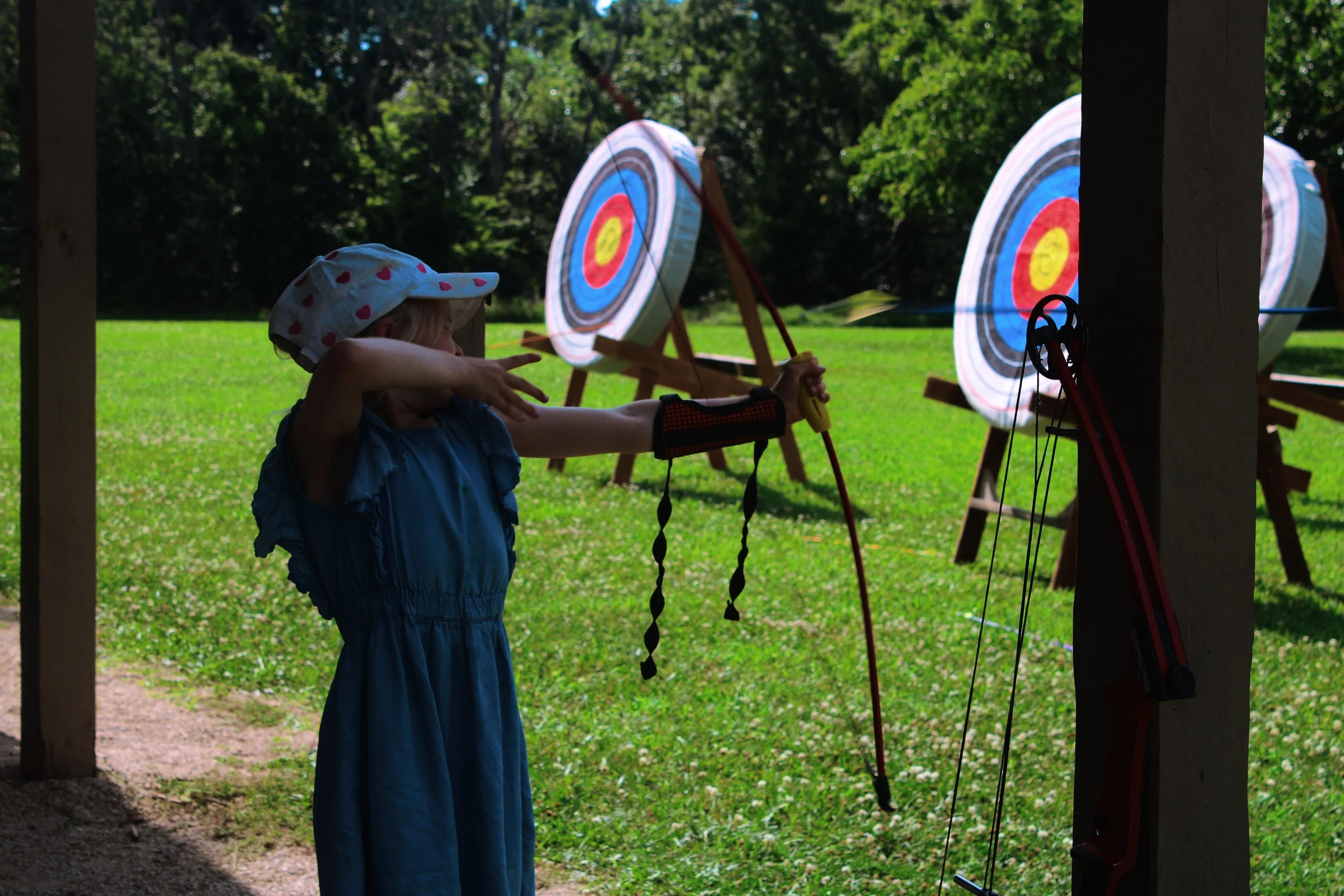 A young girl wearing a polka-dotted sun hat and blue dress is aiming at a large indoor or outdoor archery target with a bow.