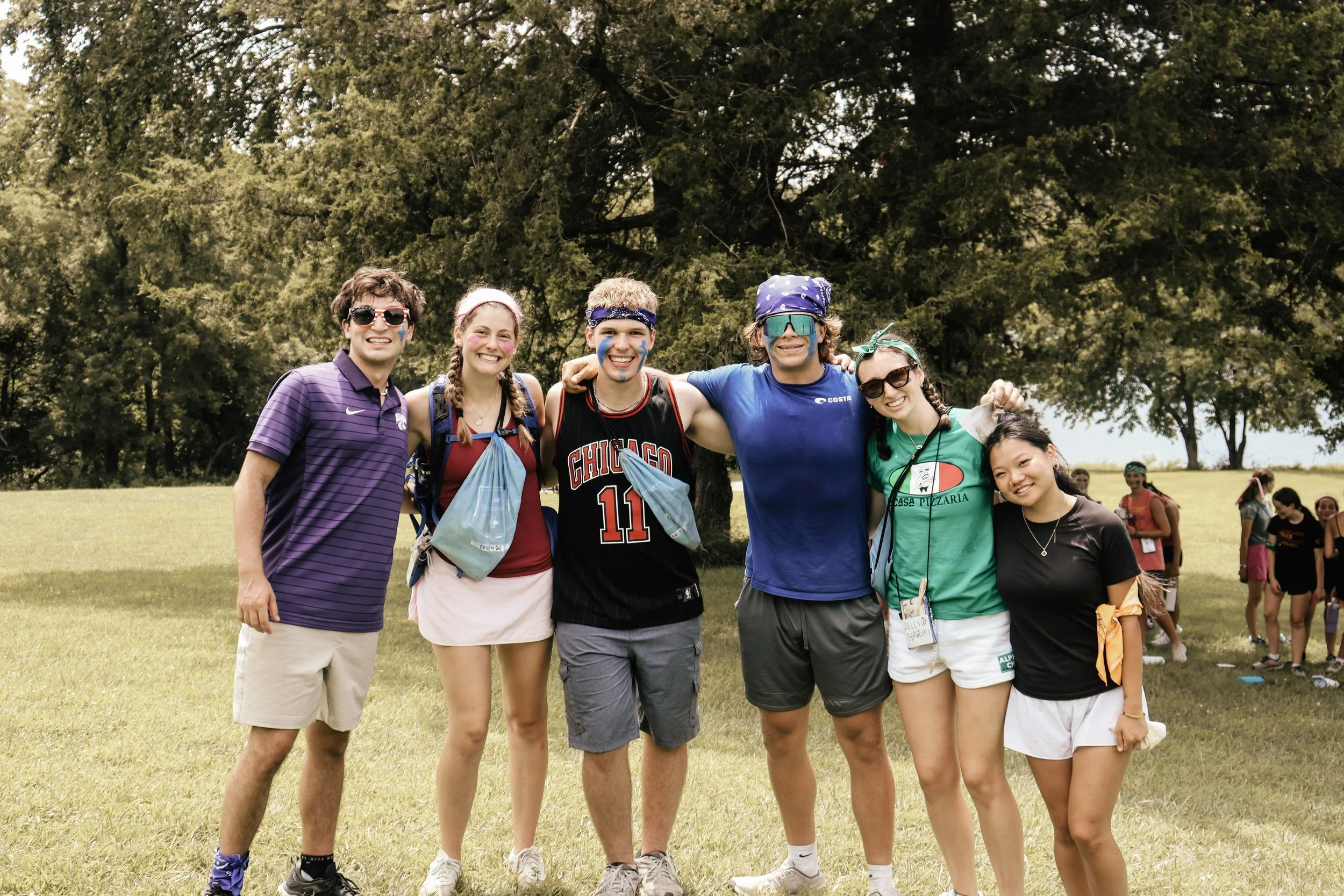 Group of six young friends smiling outdoors during daytime, standing on grass with large trees in the background.