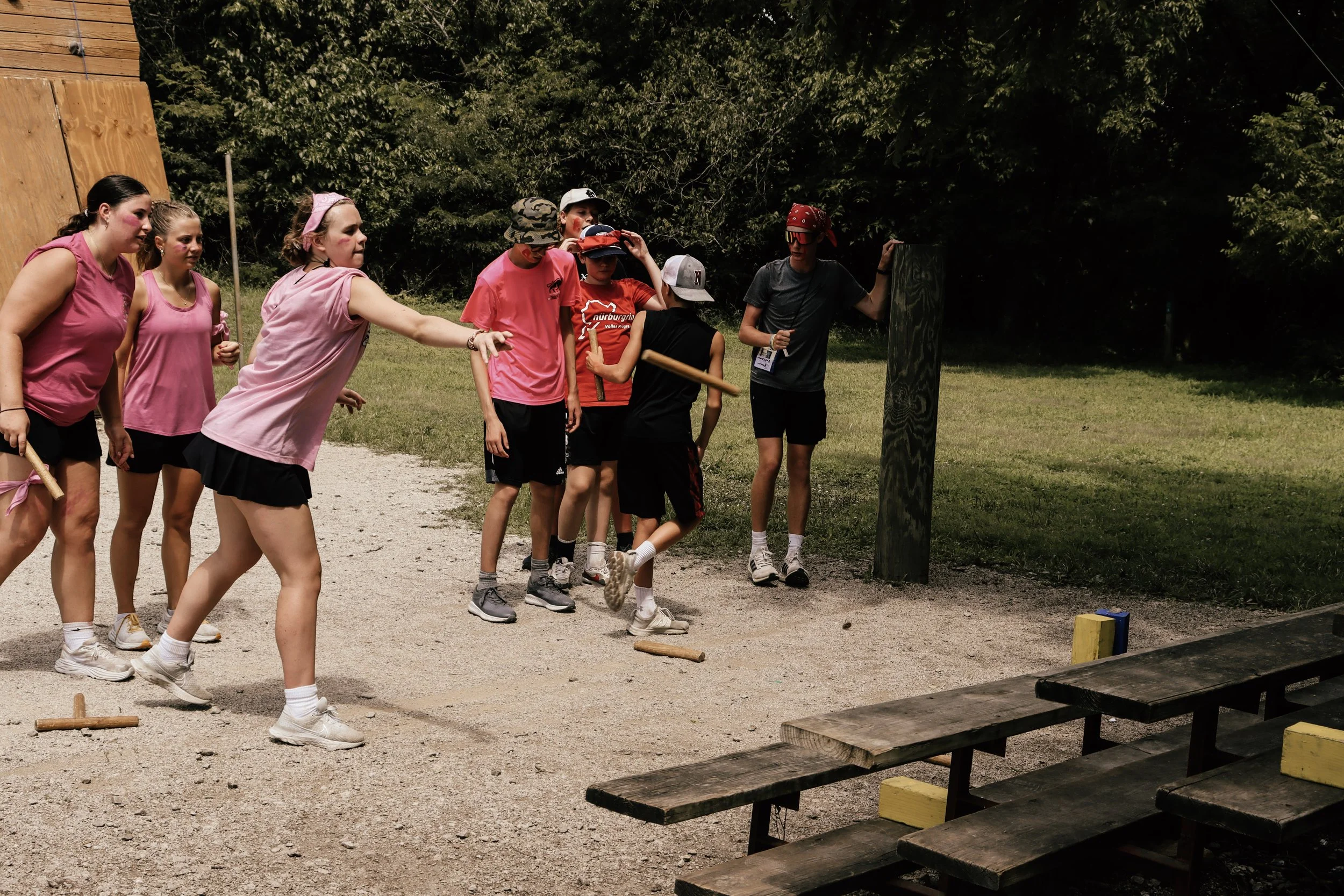 Group of children participating in an outdoor activity, some dressed in pink and casual clothes, standing on a gravel surface near a wooded area, with game or obstacle elements visible.