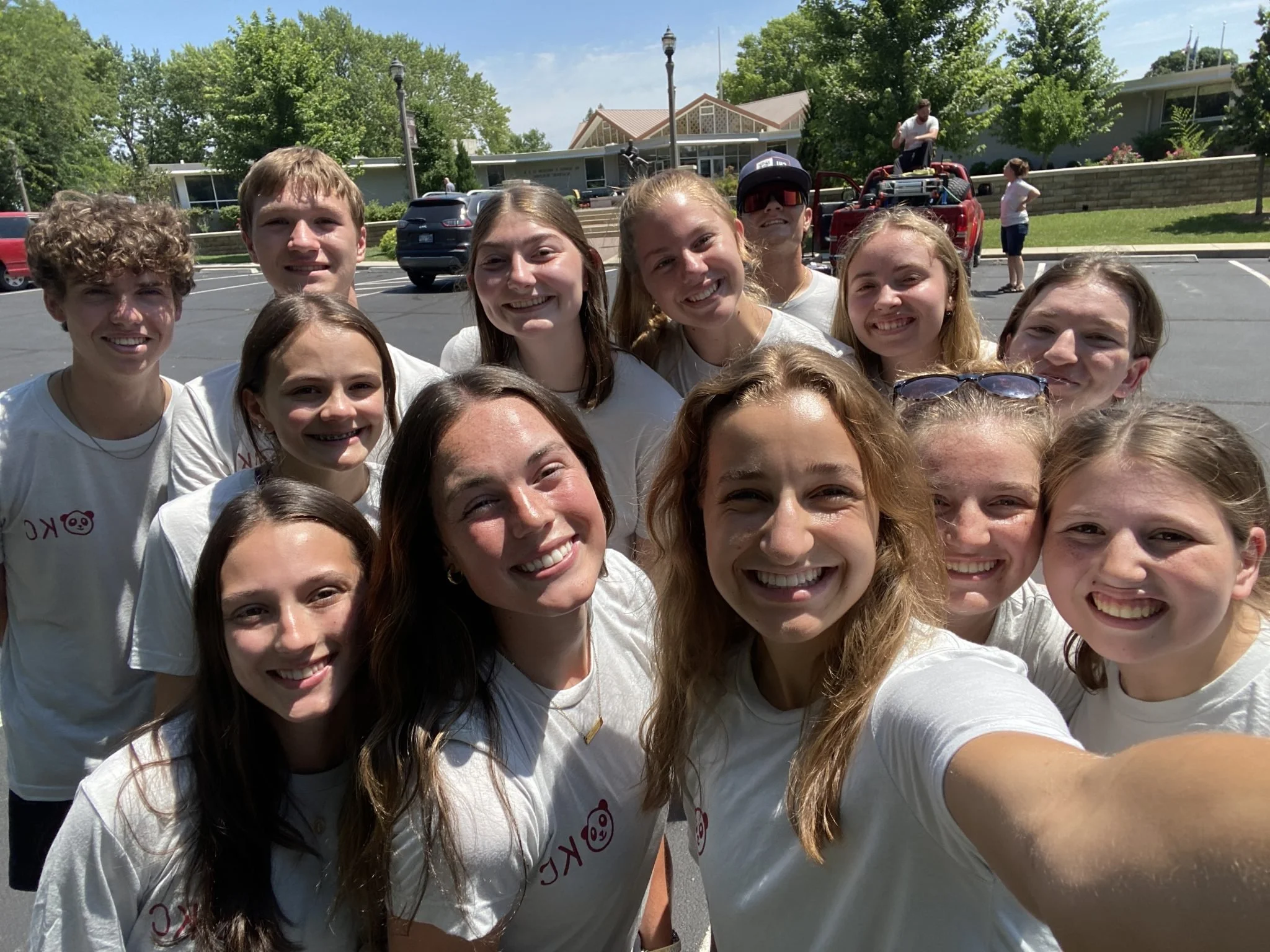 A group of smiling teenagers taking a selfie outdoors on a sunny day with trees, parked cars, and a building in the background.