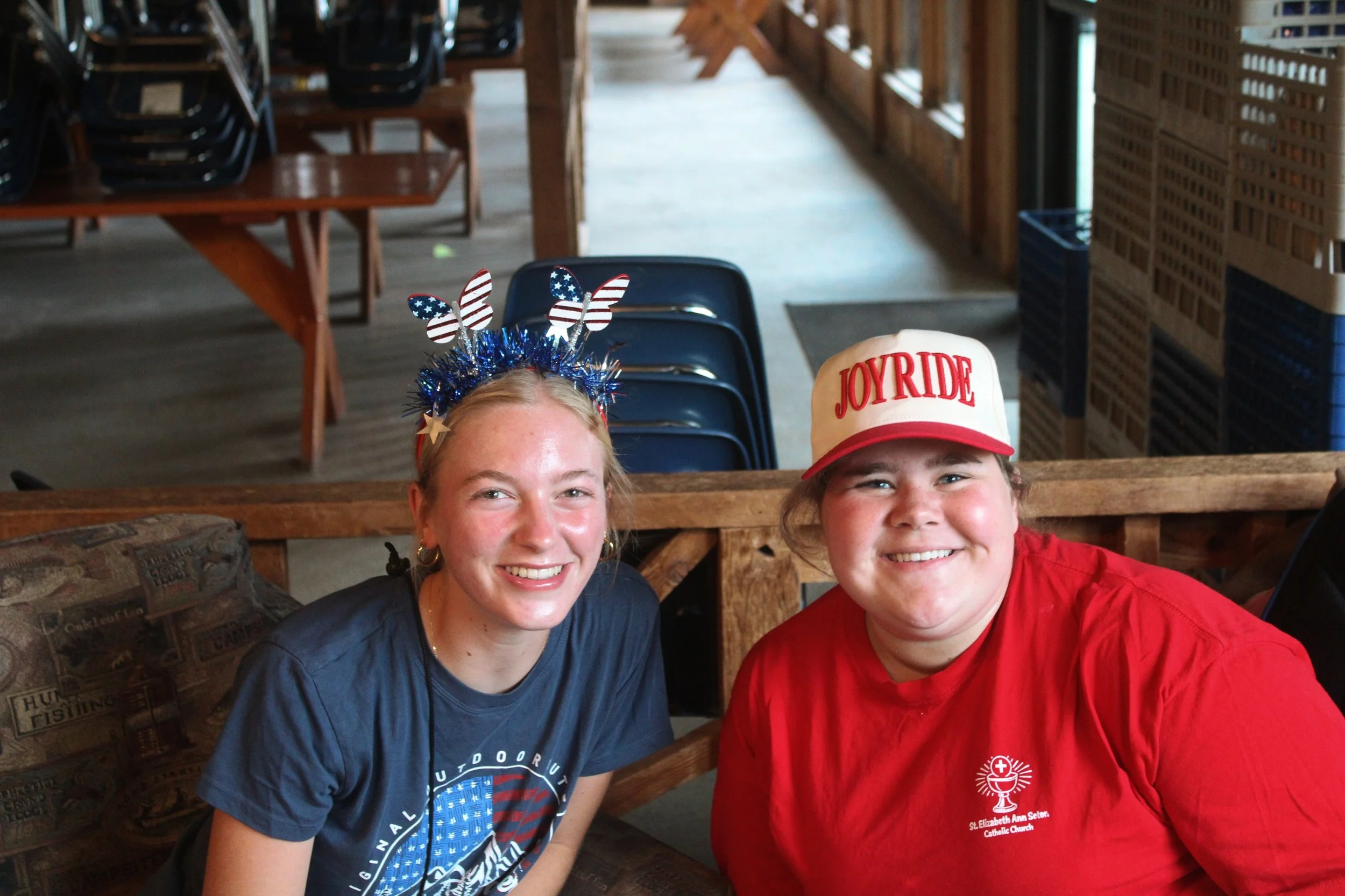 Two smiling young women sitting at a wooden table in a rustic indoor setting, wearing patriotic-themed accessories and casual clothing. One is wearing a headband with American flag patterns and a dark blue t-shirt, and the other is wearing a red base