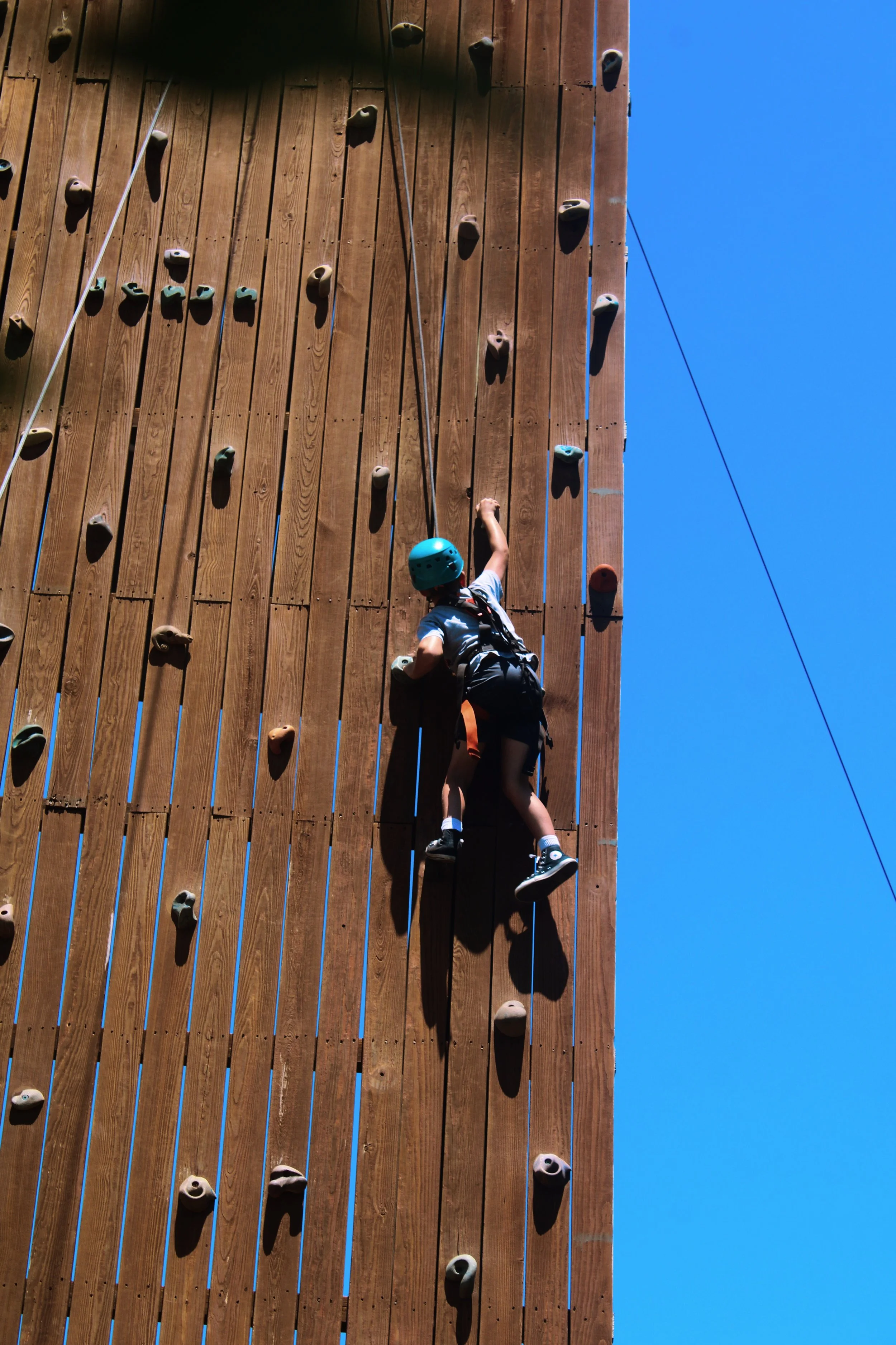 Young boy wearing a blue helmet and climbing gear scales an outdoor wooden climbing wall with colorful holds against a clear blue sky.