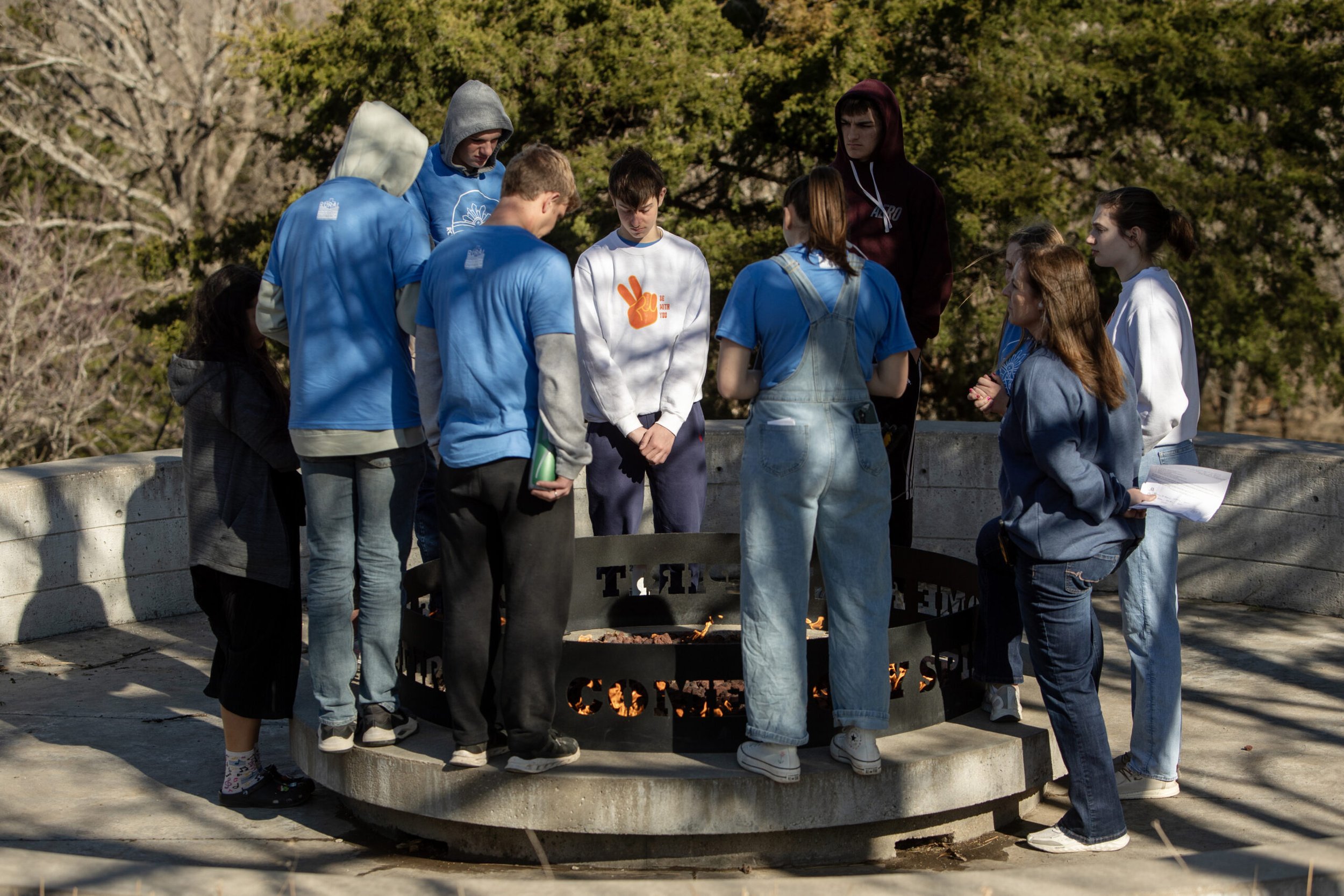 A group of young people gathered around a memorial fire pit outdoors, with some looking down and others engaged in conversation, surrounded by trees.