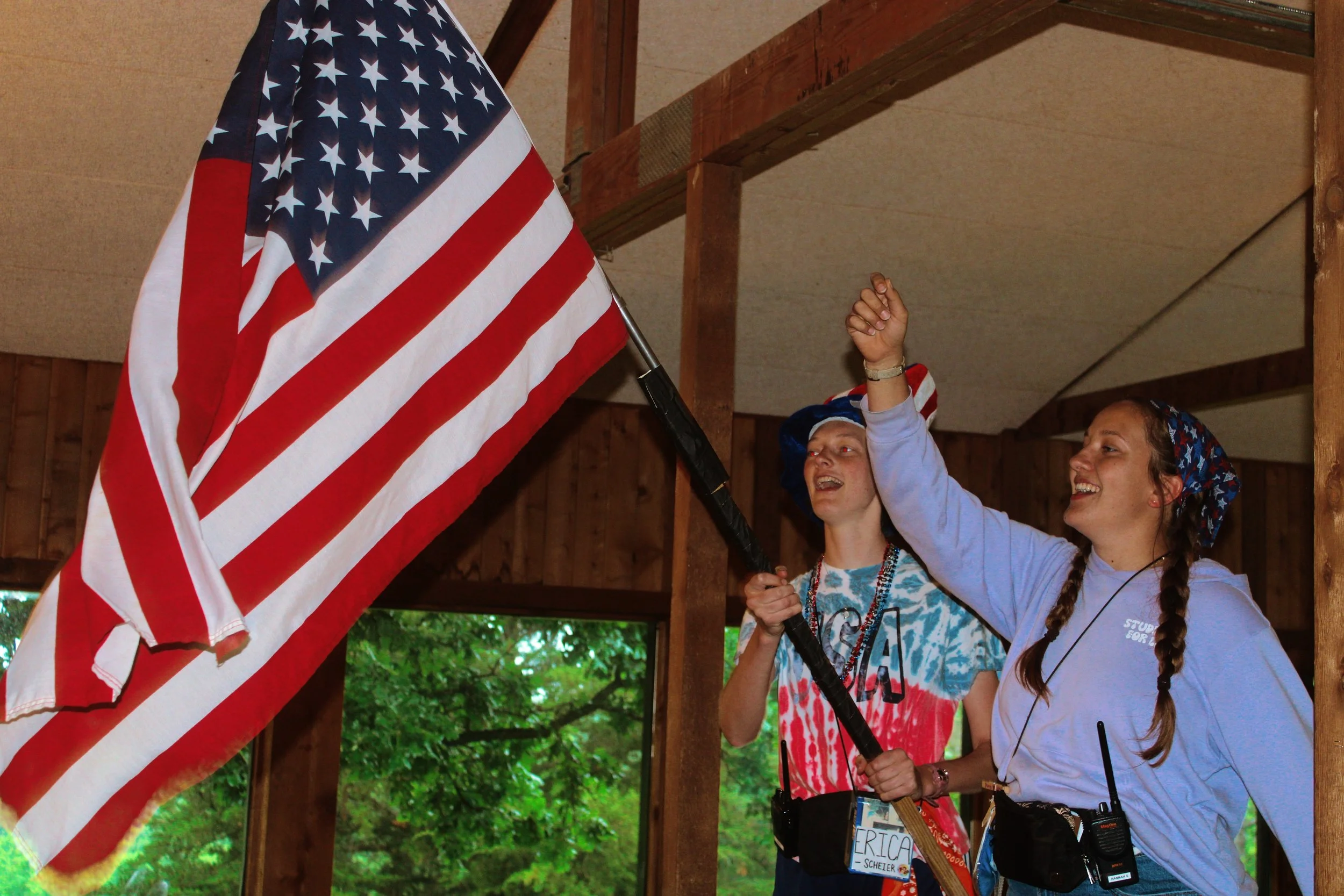 Two women are standing under a wooden structure, raising a large American flag on a flagpole. Both women are smiling, and one has her arm raised with a clenched fist. One woman is wearing a tie-dye shirt with American flag patterns, and the other is 