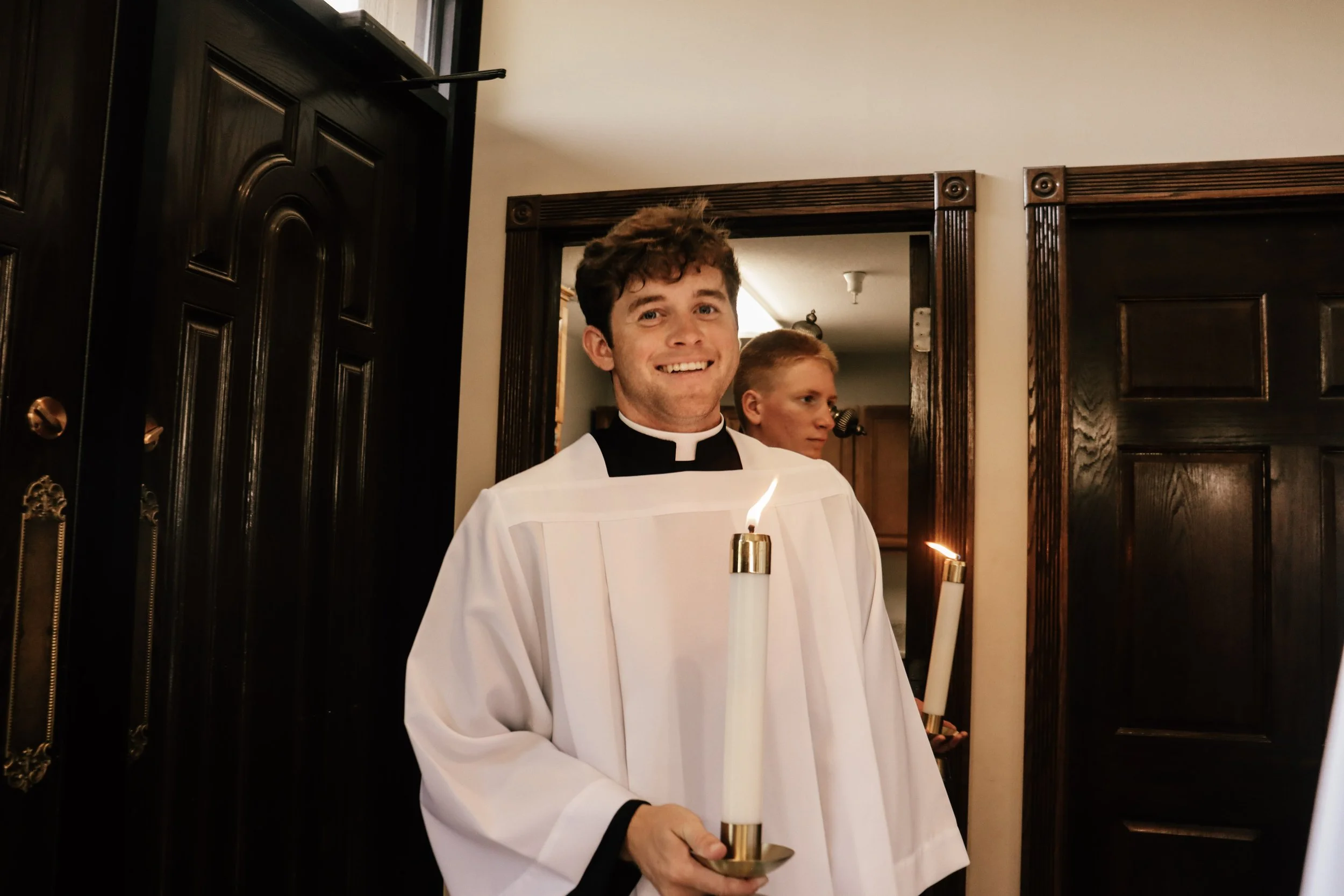 A young man in a church or religious setting wearing a white robe with a black collar, holding a brass candle holder with a lit candle. Behind him, another young man in similar attire. The room has dark wooden trim and doors.