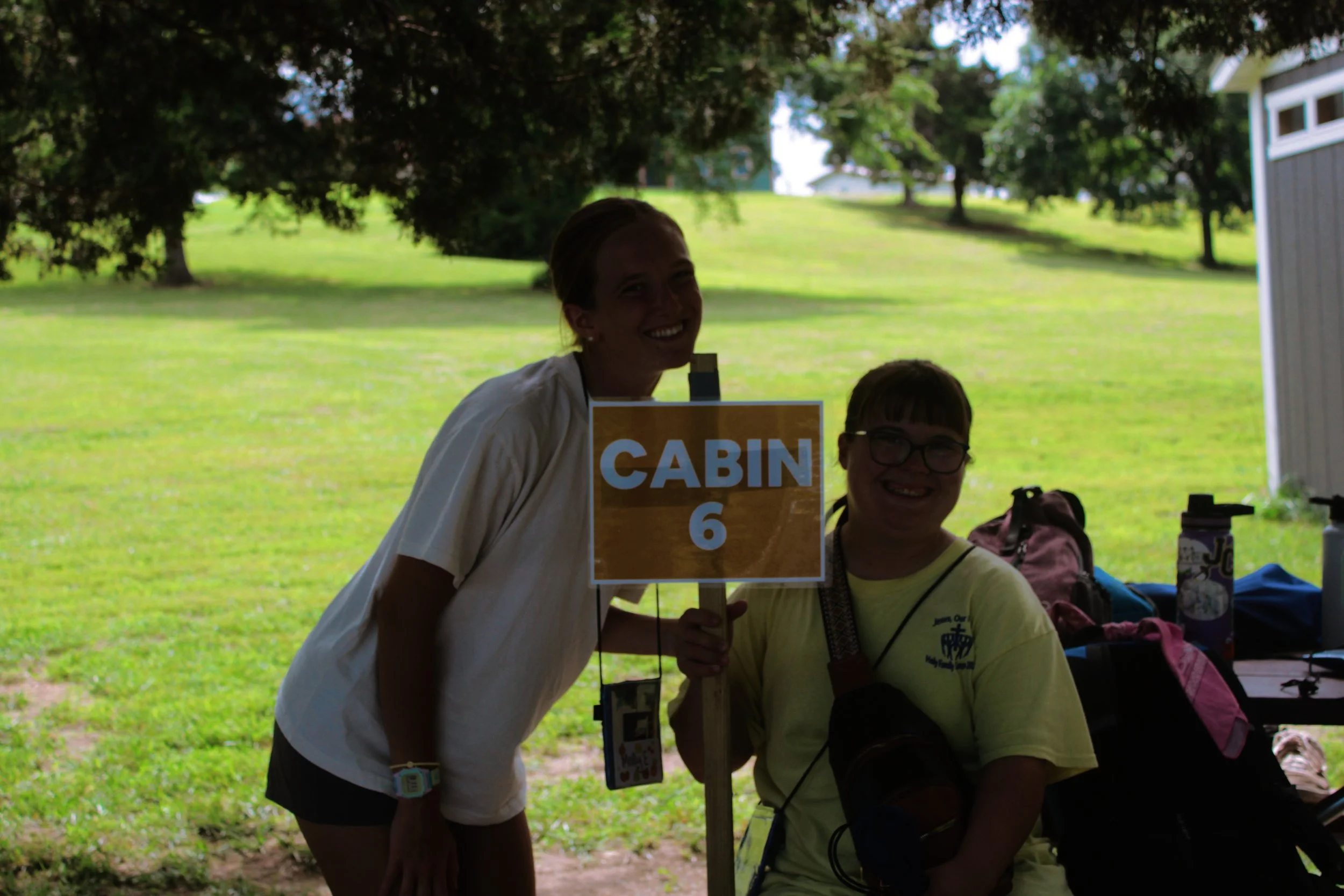 Two women holding a sign that says 'CABIN 6' outdoors in a park or outdoor area with trees in the background.