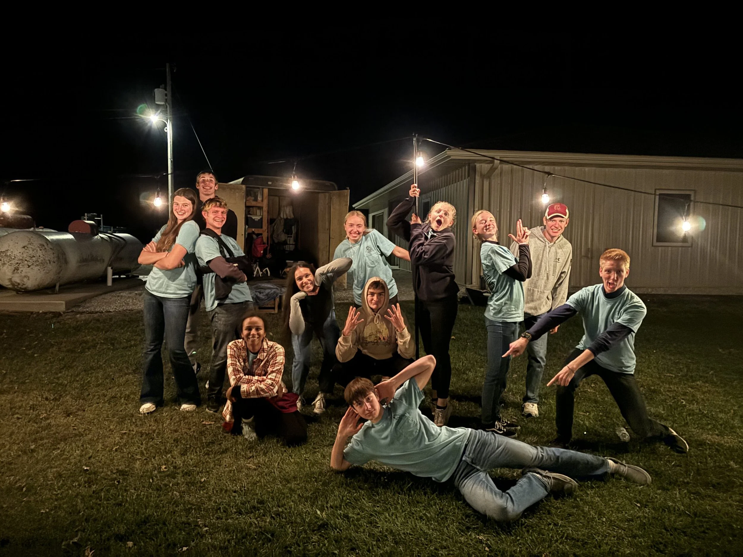 A group of smiling children and teenagers posing outdoors at night, some sitting and some standing, with string lights and a building in the background.