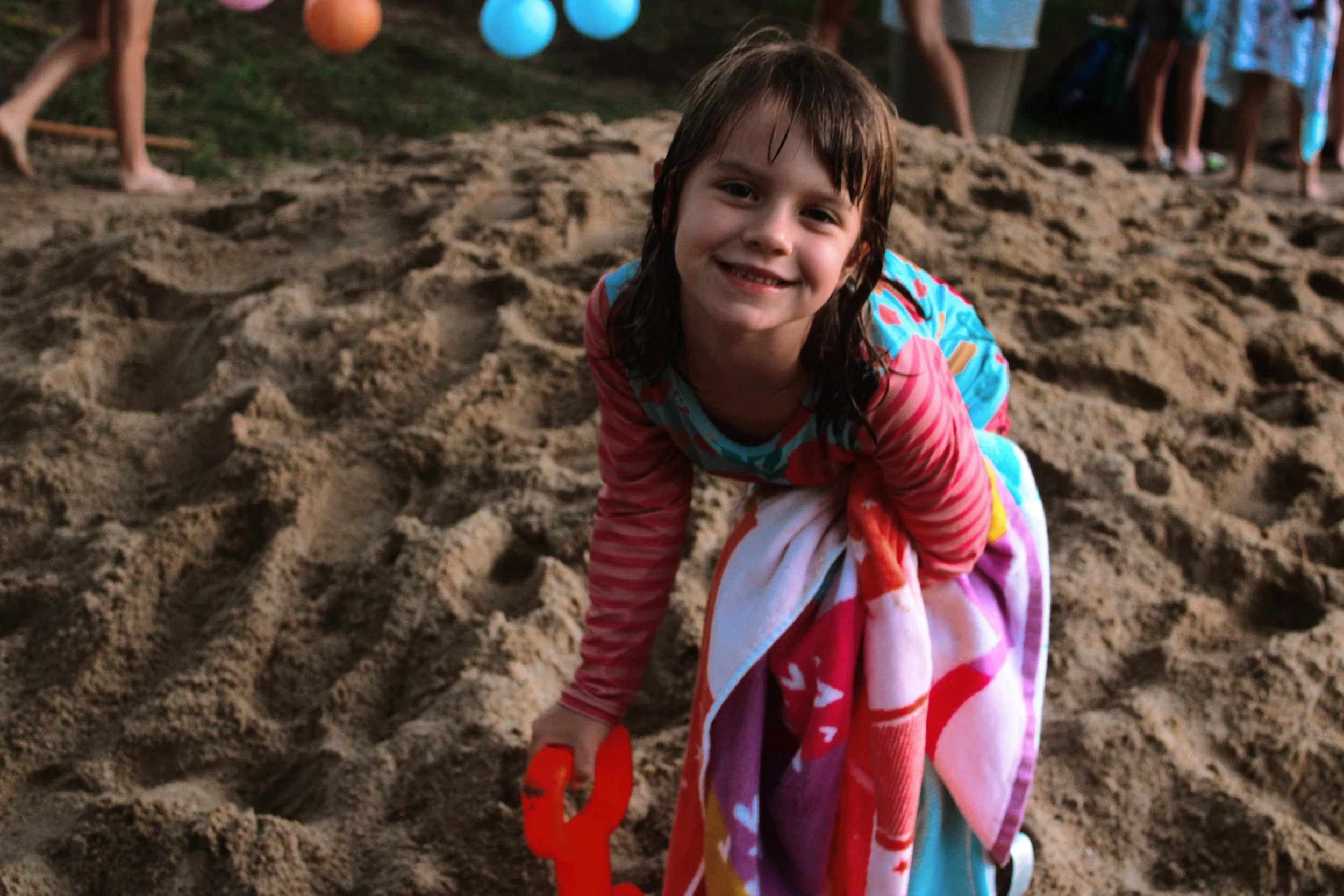 A smiling girl with wet hair, wearing a colorful beach towel, is kneeling in the sand and holding a small orange shovel. In the background, there are children and colorful beach balls.