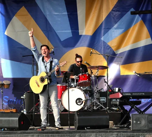 Two musicians performing on stage, one singing with a guitar and hand raised, the other playing drums, with colorful sunburst backdrop.