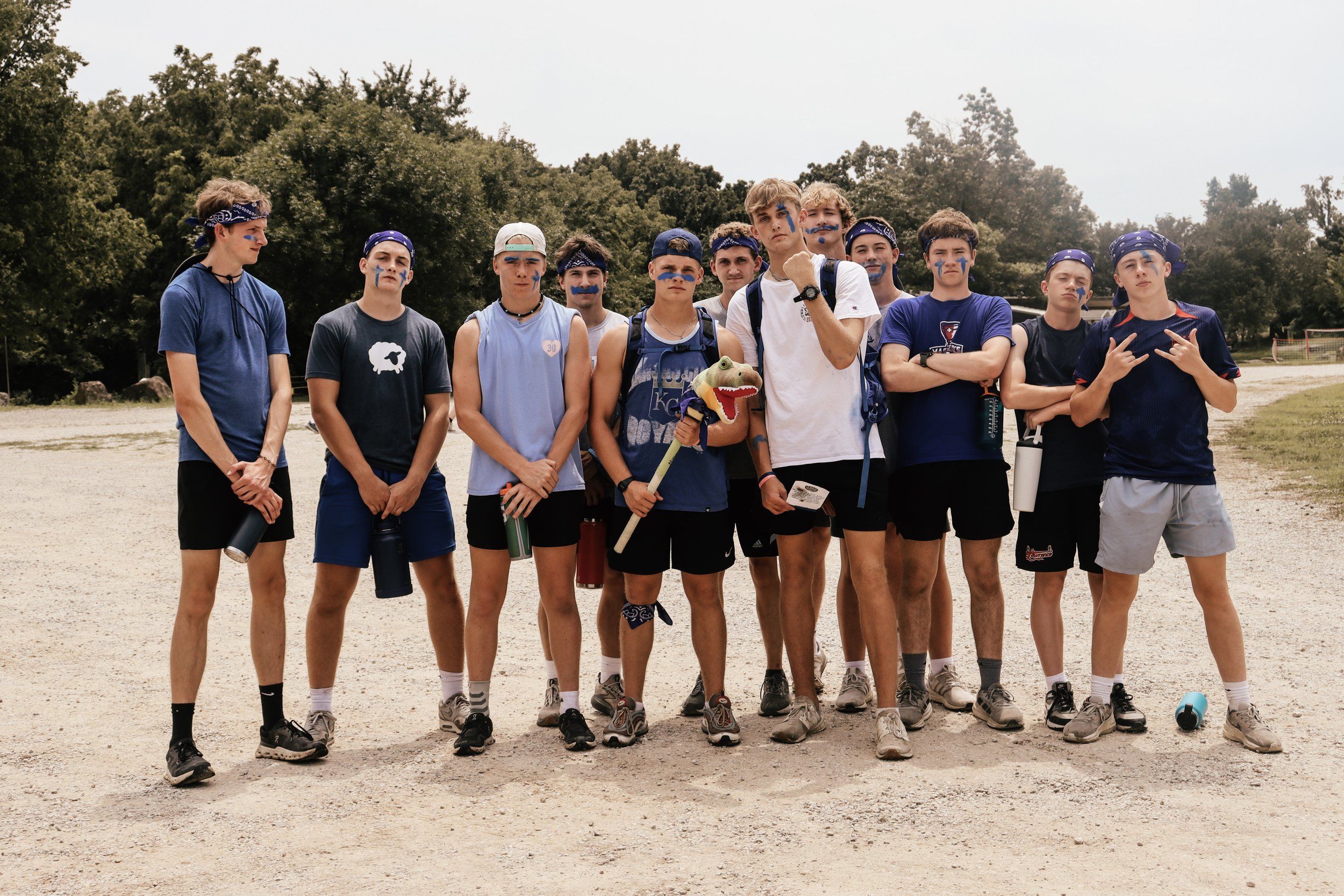 A group of young boys dressed in athletic clothing and face paint, standing outdoors on a dirt field, some with backpacks and a dog puppet, with trees in the background.