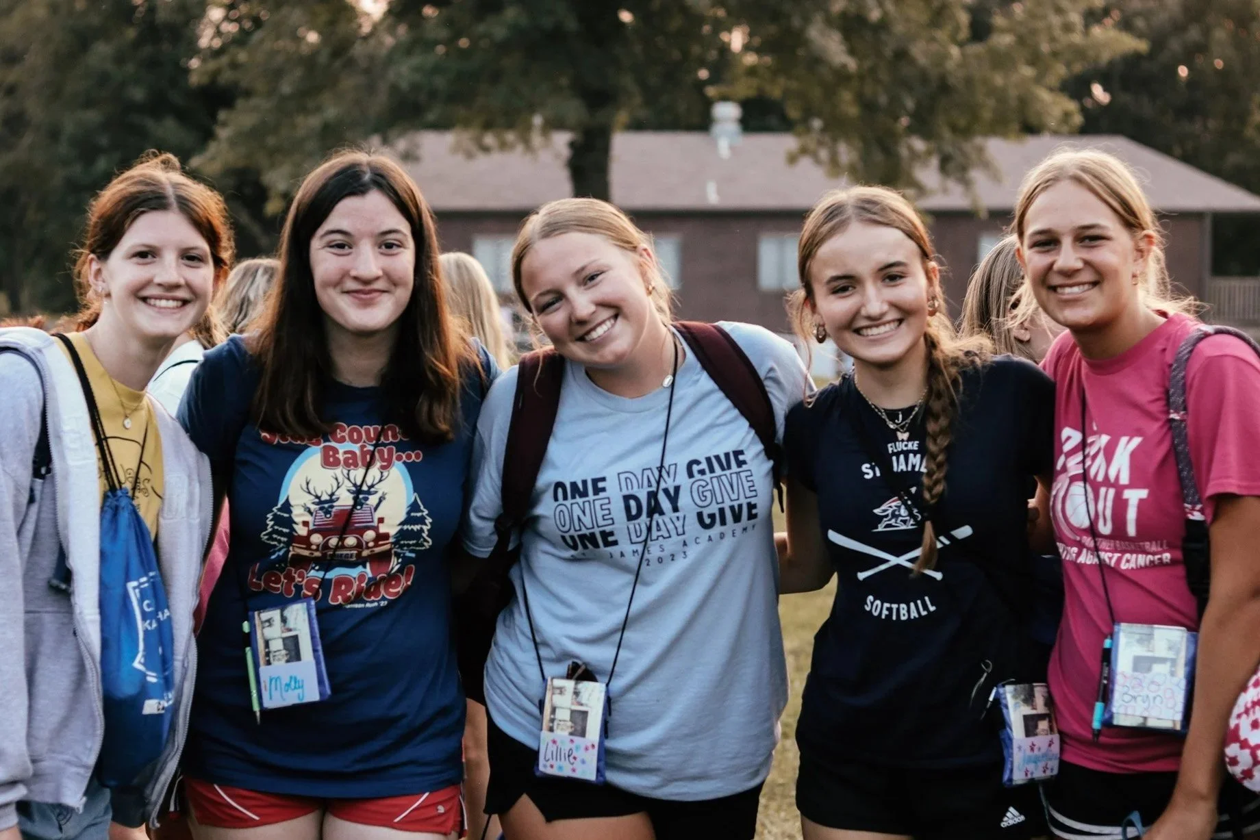 A group of five young women smiling and standing close together outdoors during daytime, with a house and trees in the background. They are wearing casual sports t-shirts and shorts, each with a name tag and backpack.